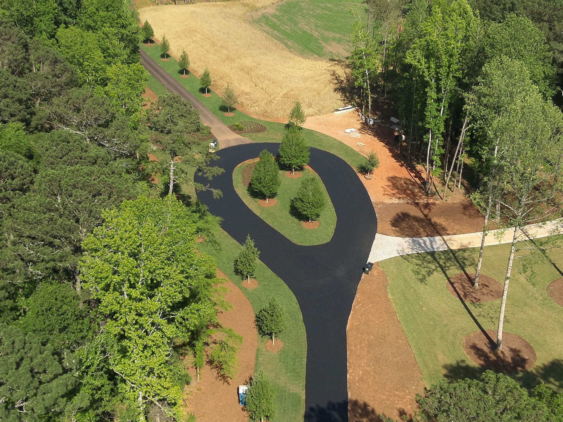 Aerial view of a black asphalt driveway with a circular turnaround, lined with trees and grass, in a wooded area.