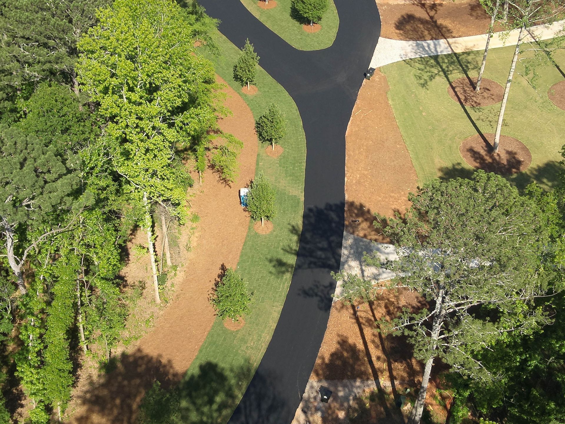 Aerial view of winding black pathway through green grass and brown mulch with trees.