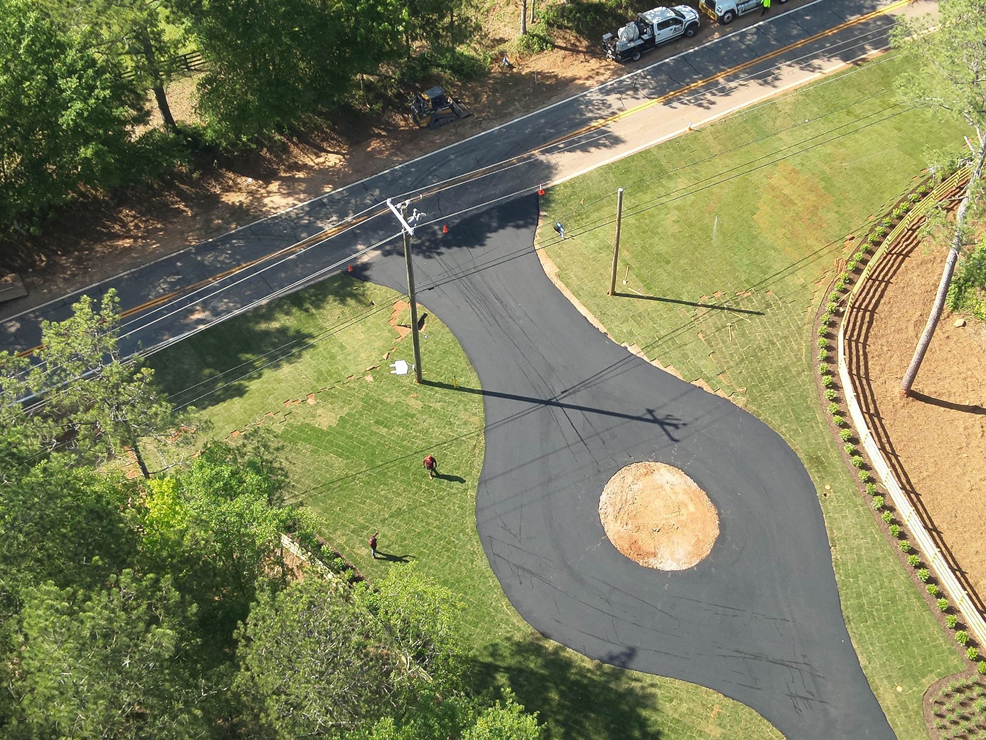 Aerial view of new asphalt driveway leading to road, with a grassy roundabout. Green trees and grass surround.