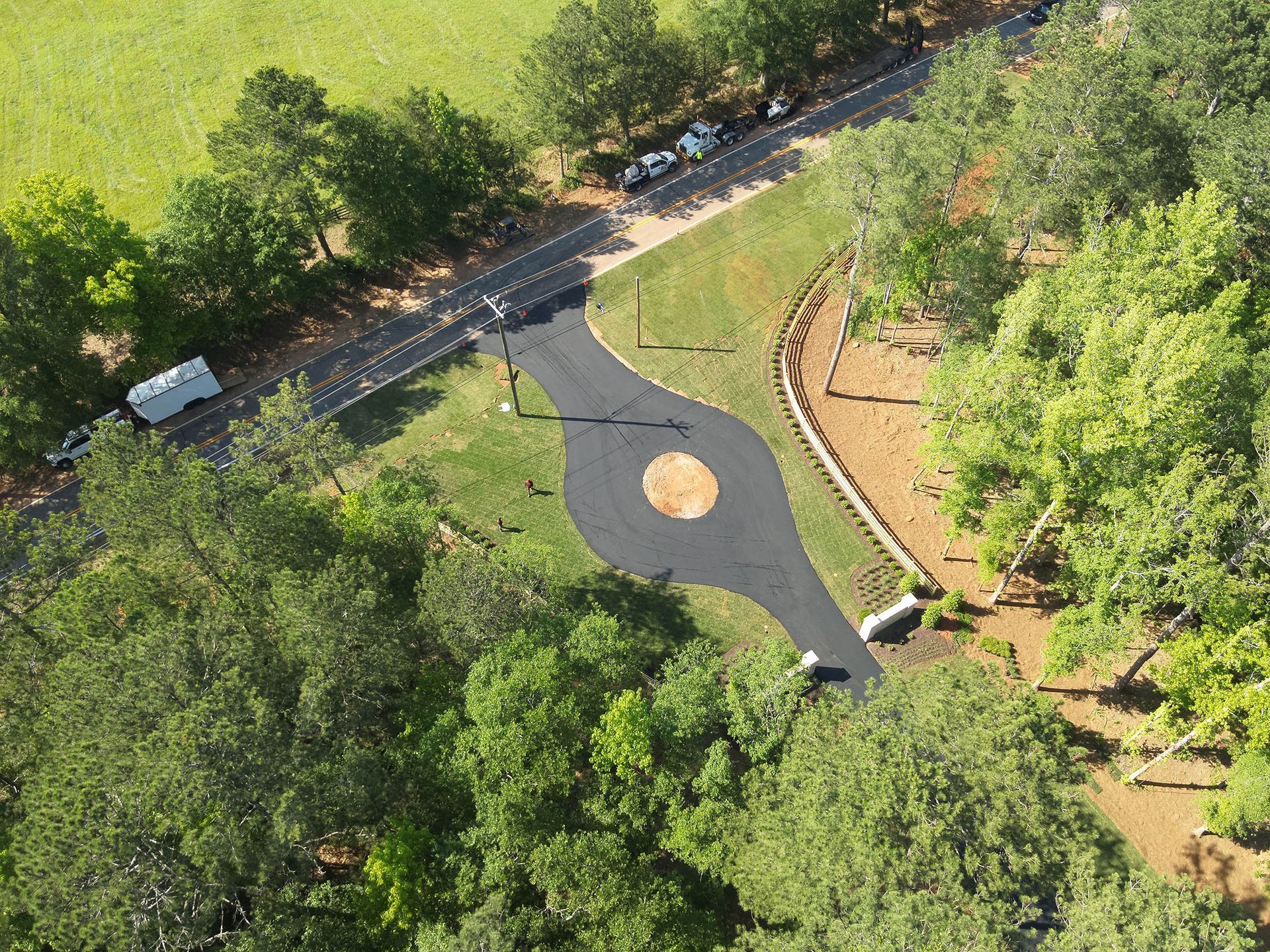 Aerial view of a paved roundabout on a road, surrounded by trees and a green field.