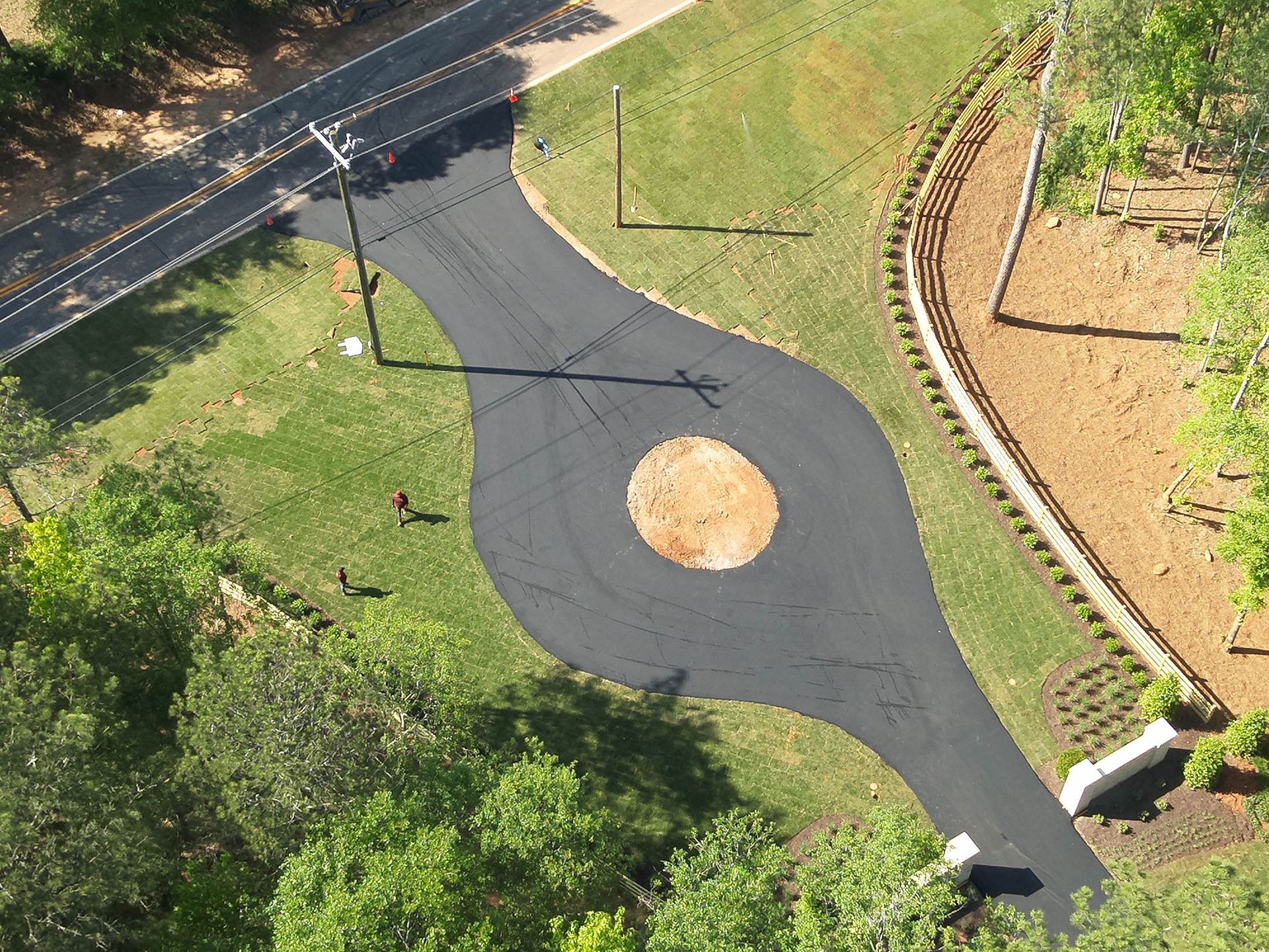 Overhead view of a black asphalt driveway with a dirt island. Green lawn surrounds it, and a road is visible.