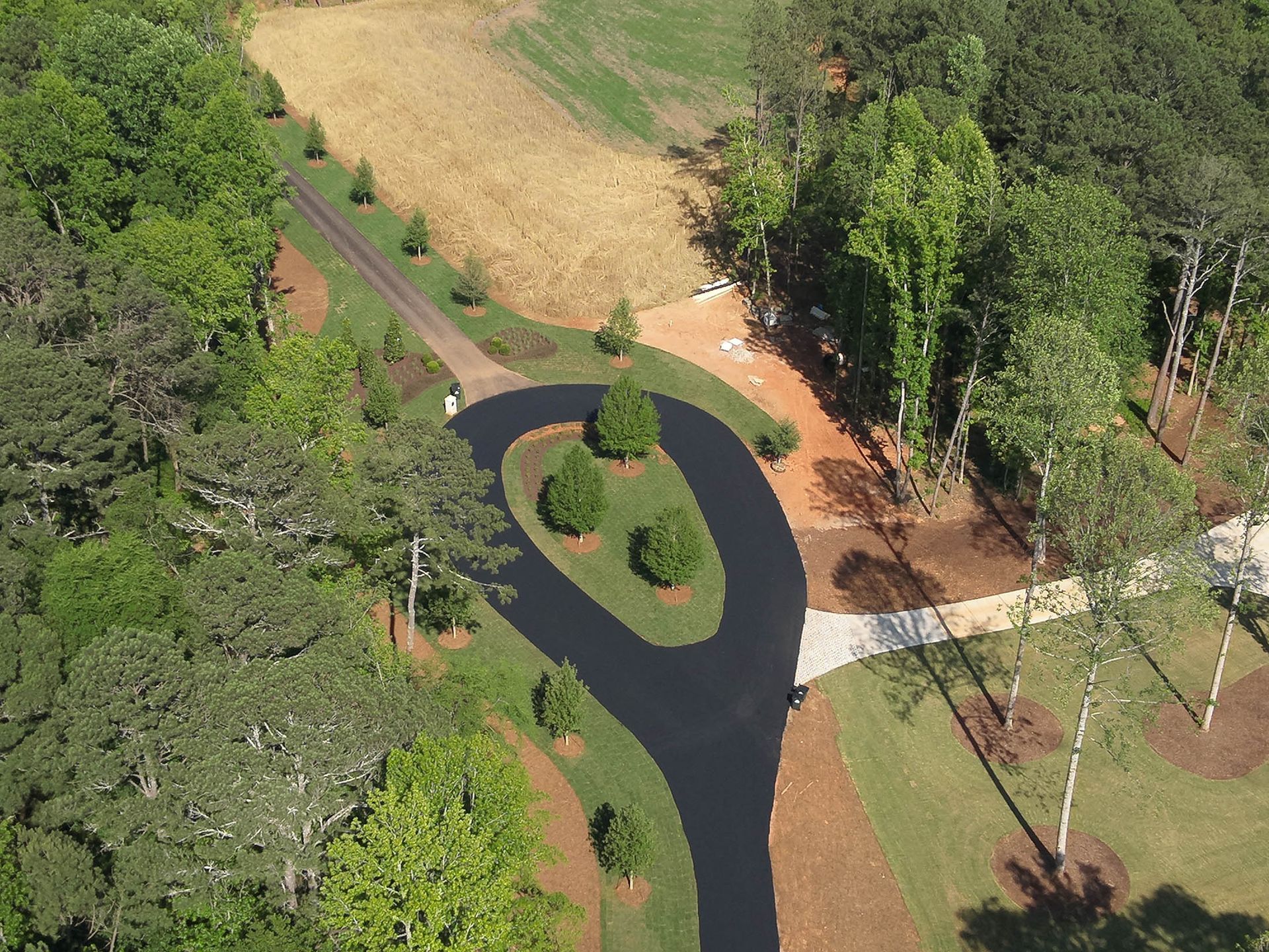 Aerial view of a paved driveway with a circular turnaround, surrounded by trees, grass, and a field.