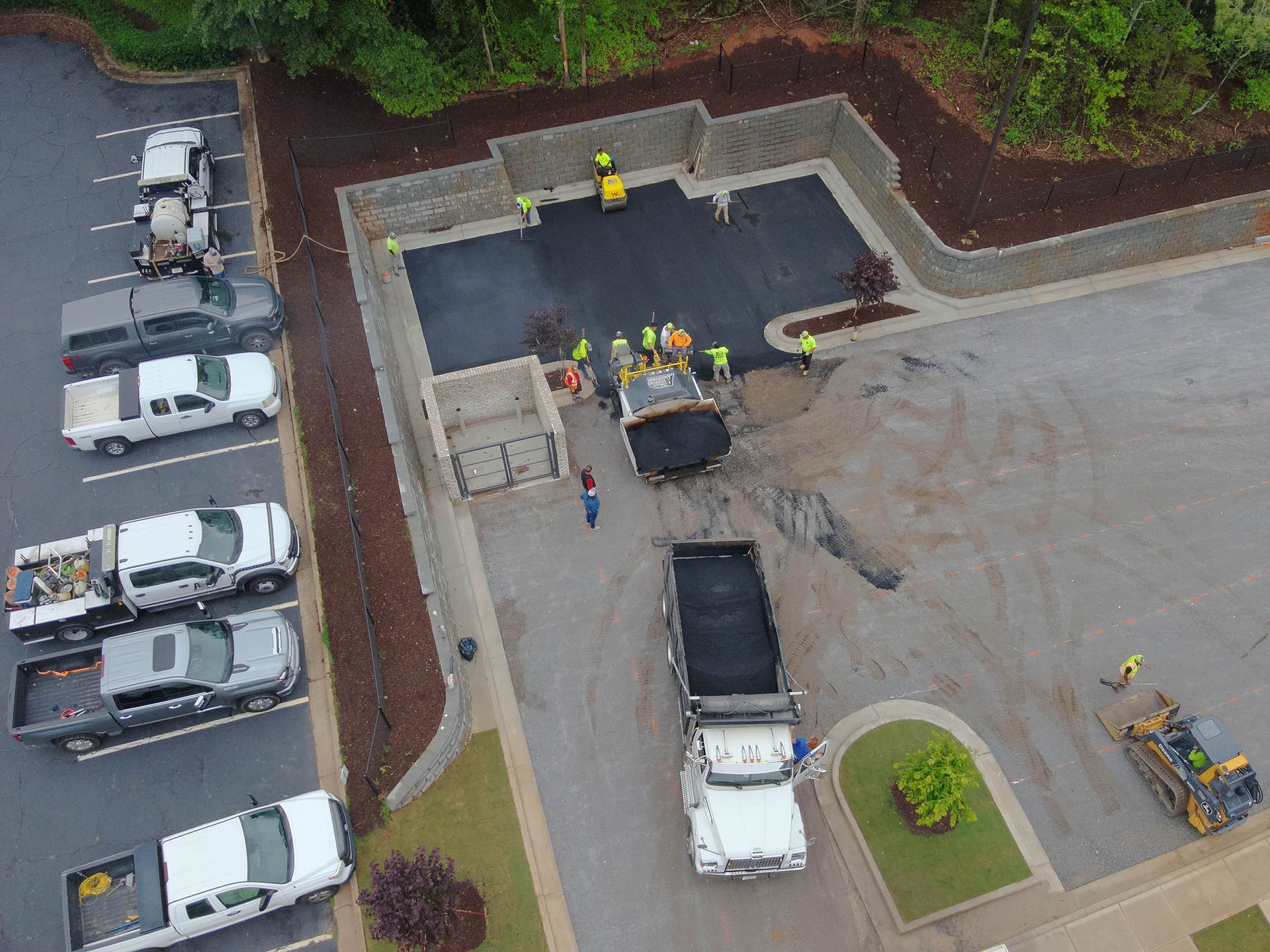 Asphalt paving crew at work, laying blacktop in a parking area, surrounded by trucks and landscaping.