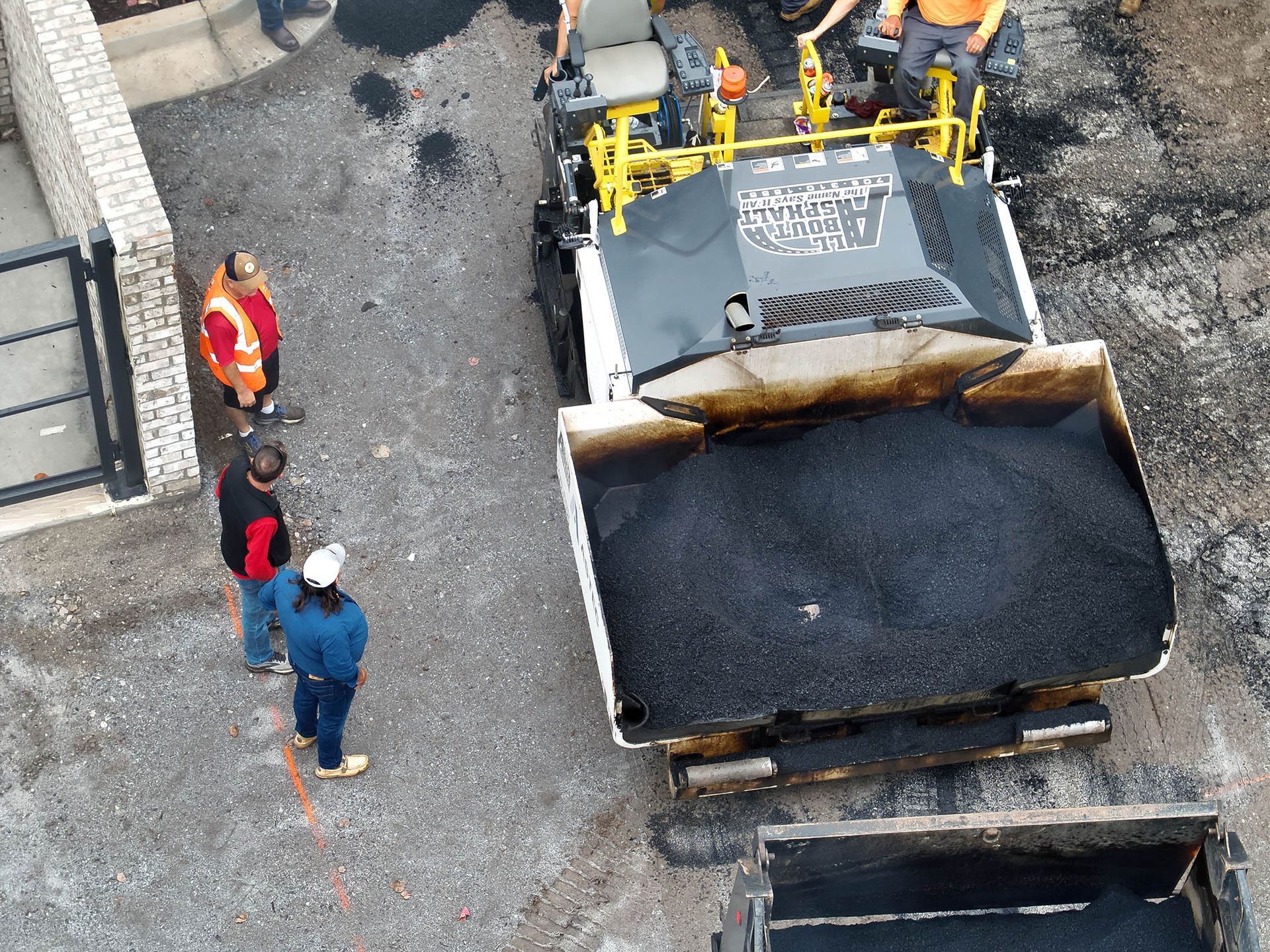 Asphalt paving crew at work, a truck dumps fresh asphalt into a paver. Workers observe.