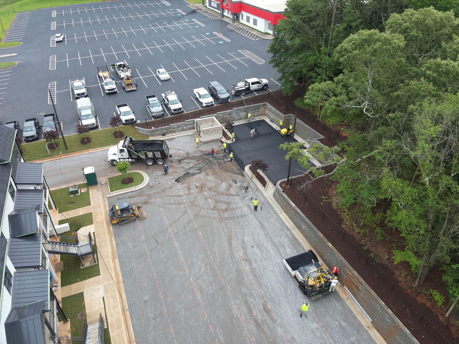Construction site with paving crew, dump trucks, and heavy machinery, near a parking lot and building.