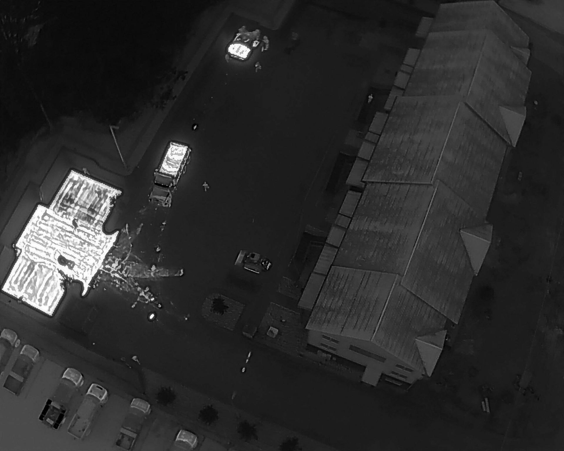 Overhead grayscale view of a building with cars, a parking area, and a smaller structure illuminated.
