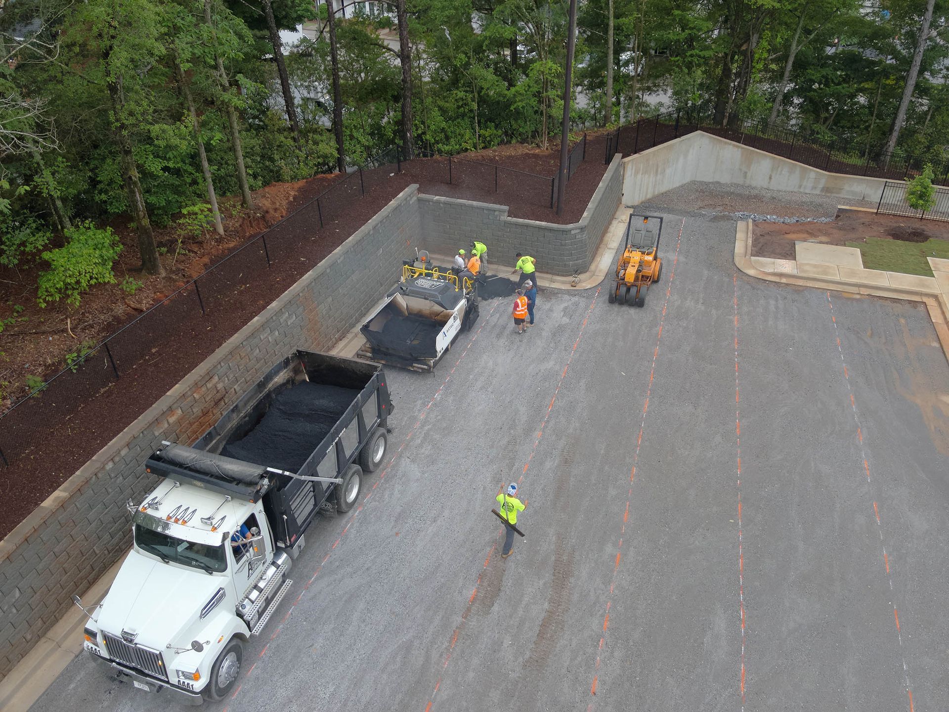 Asphalt paving operation with dump truck, workers, and machinery near a retaining wall.