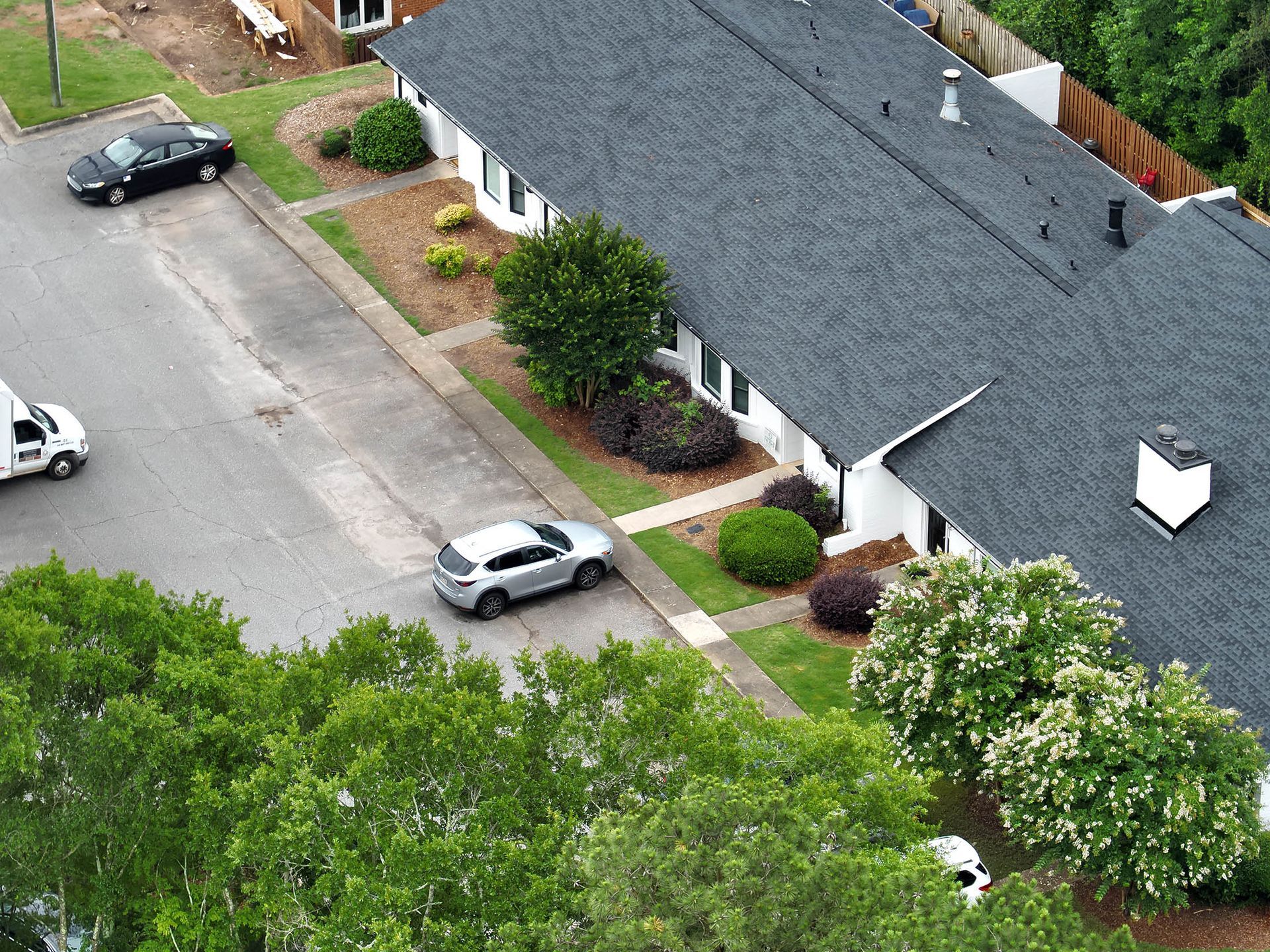 Overhead view of a white building with black roof, cars parked along the street, and greenery.