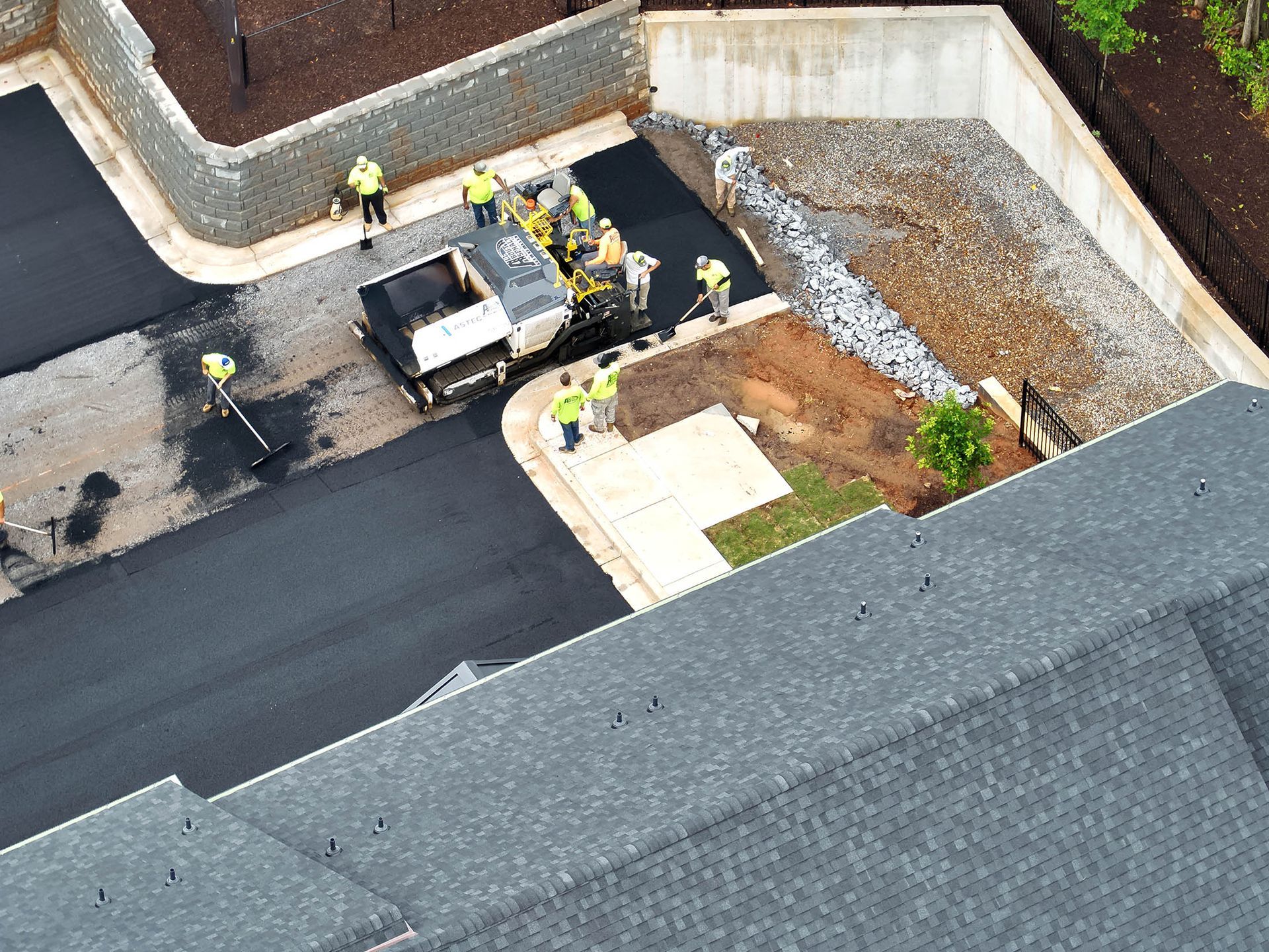 Asphalt paving crew at work, laying pavement in a residential area. Men in yellow vests operate machinery and spread asphalt.