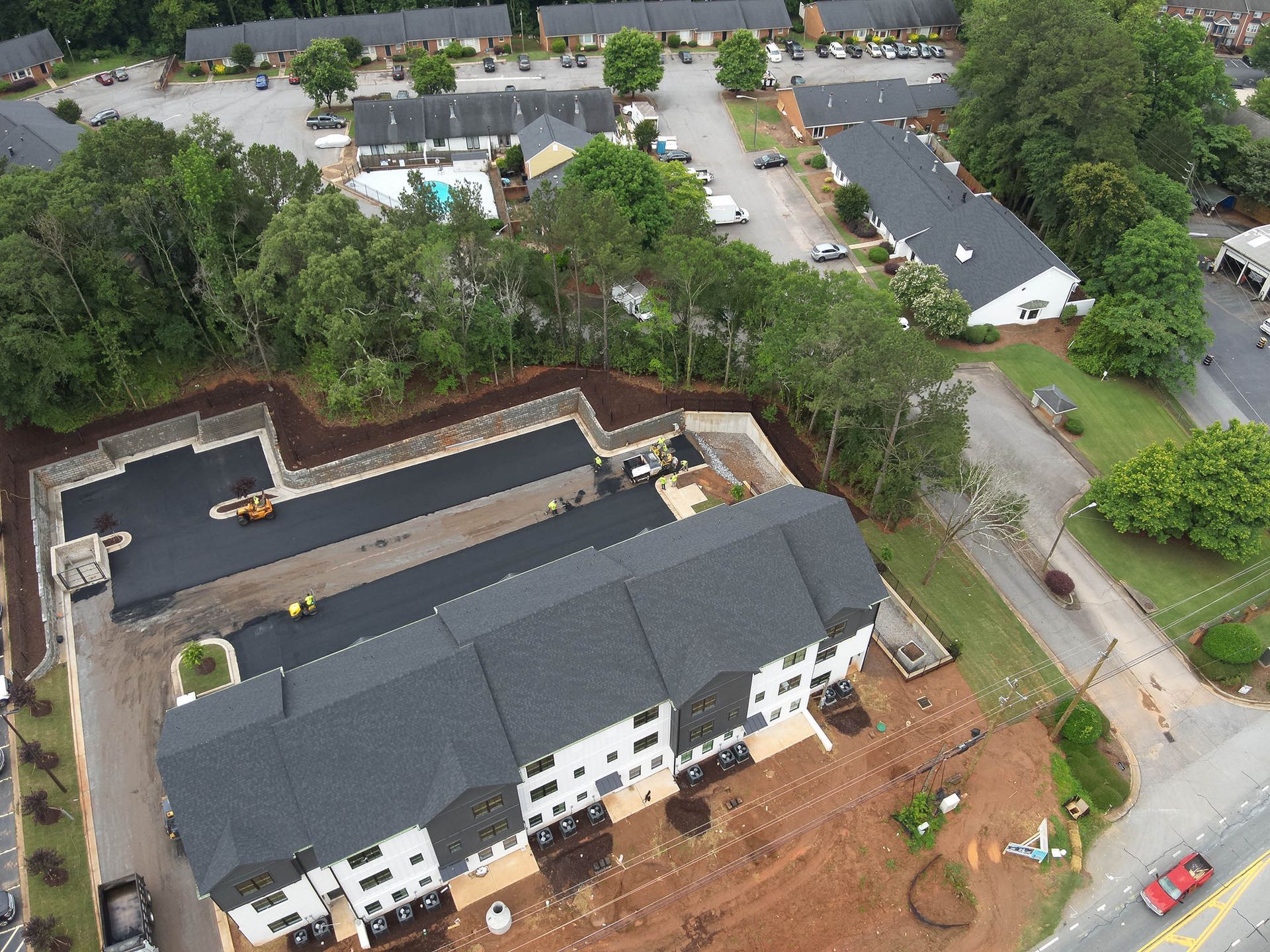 Aerial view of multi-story apartment buildings with dark gray roofs, surrounded by trees and newly paved driveways.