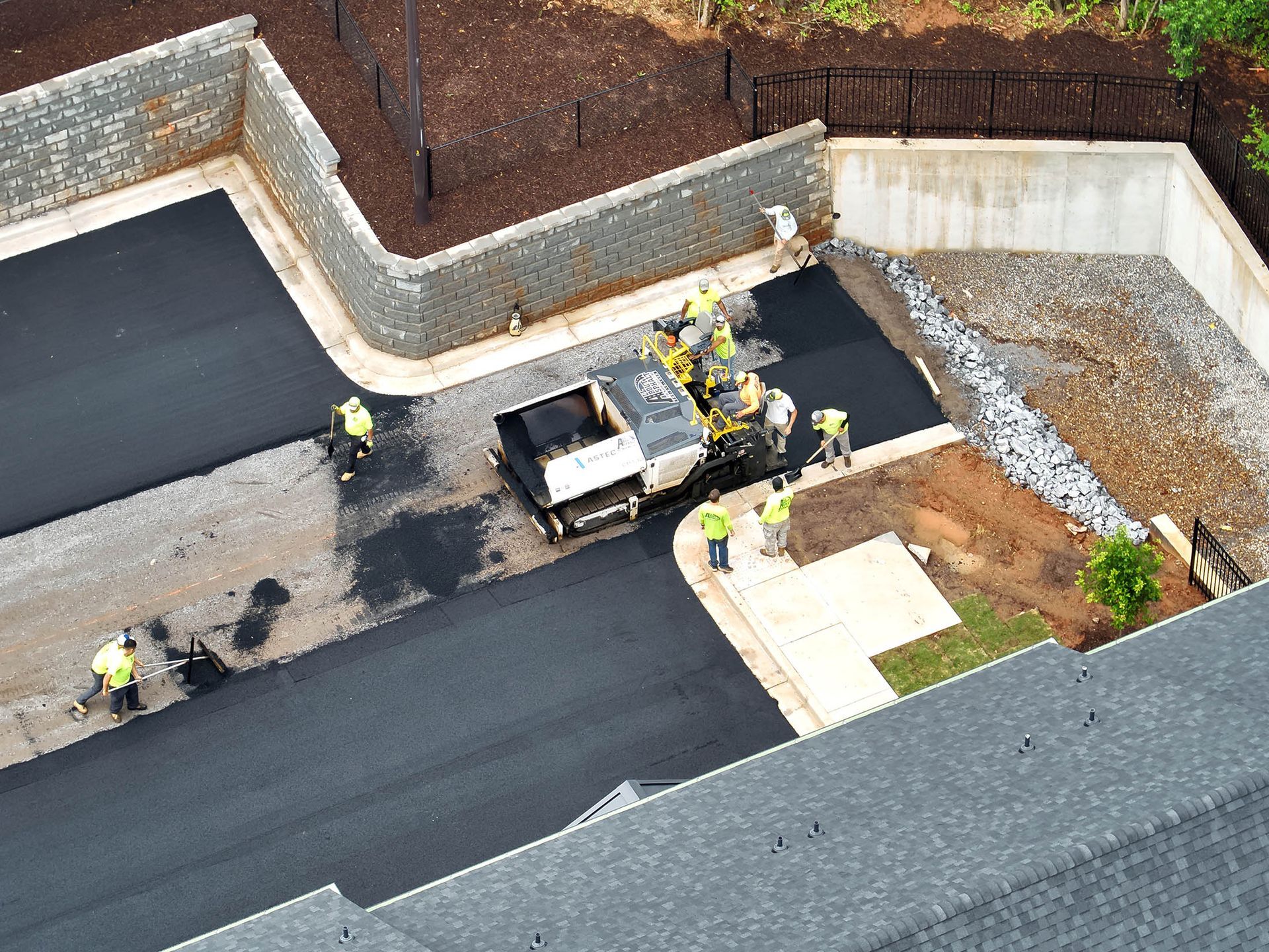 Asphalt paving crew at work, laying blacktop in a parking area, surrounded by retaining walls and landscaping.