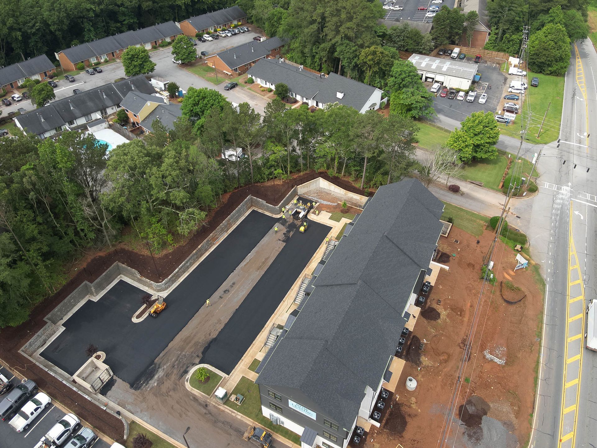 Aerial view of a construction site. Asphalt paving for a building's parking lot, with existing townhouses and road in the background.