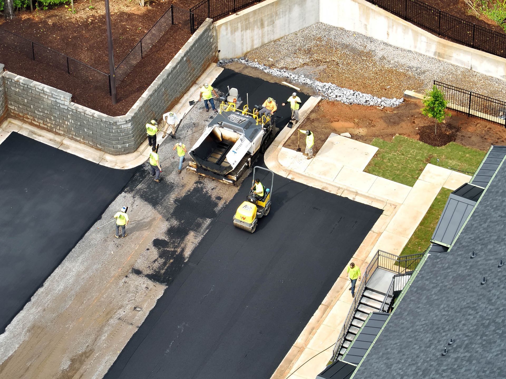 Workers paving a parking lot with asphalt. A machine lays the black surface, while others assist and a roller compacts it.