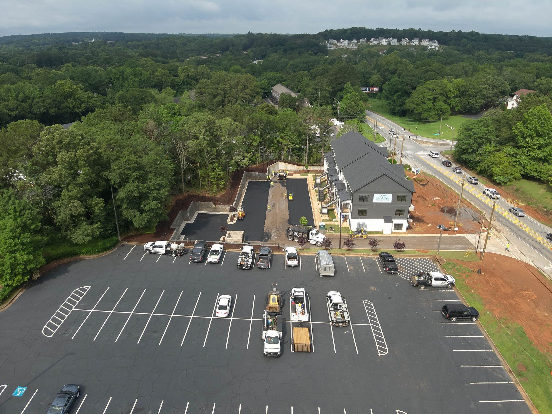 Aerial view of asphalt parking lot with construction vehicles and new building along a road, bordered by trees.