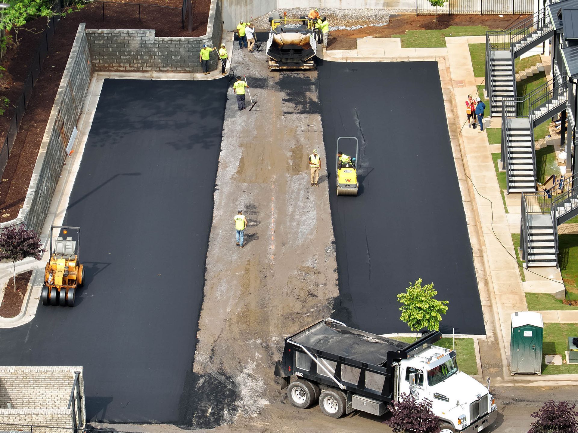Asphalt paving in progress. Workers and machinery laying blacktop in a parking area, dump truck visible.