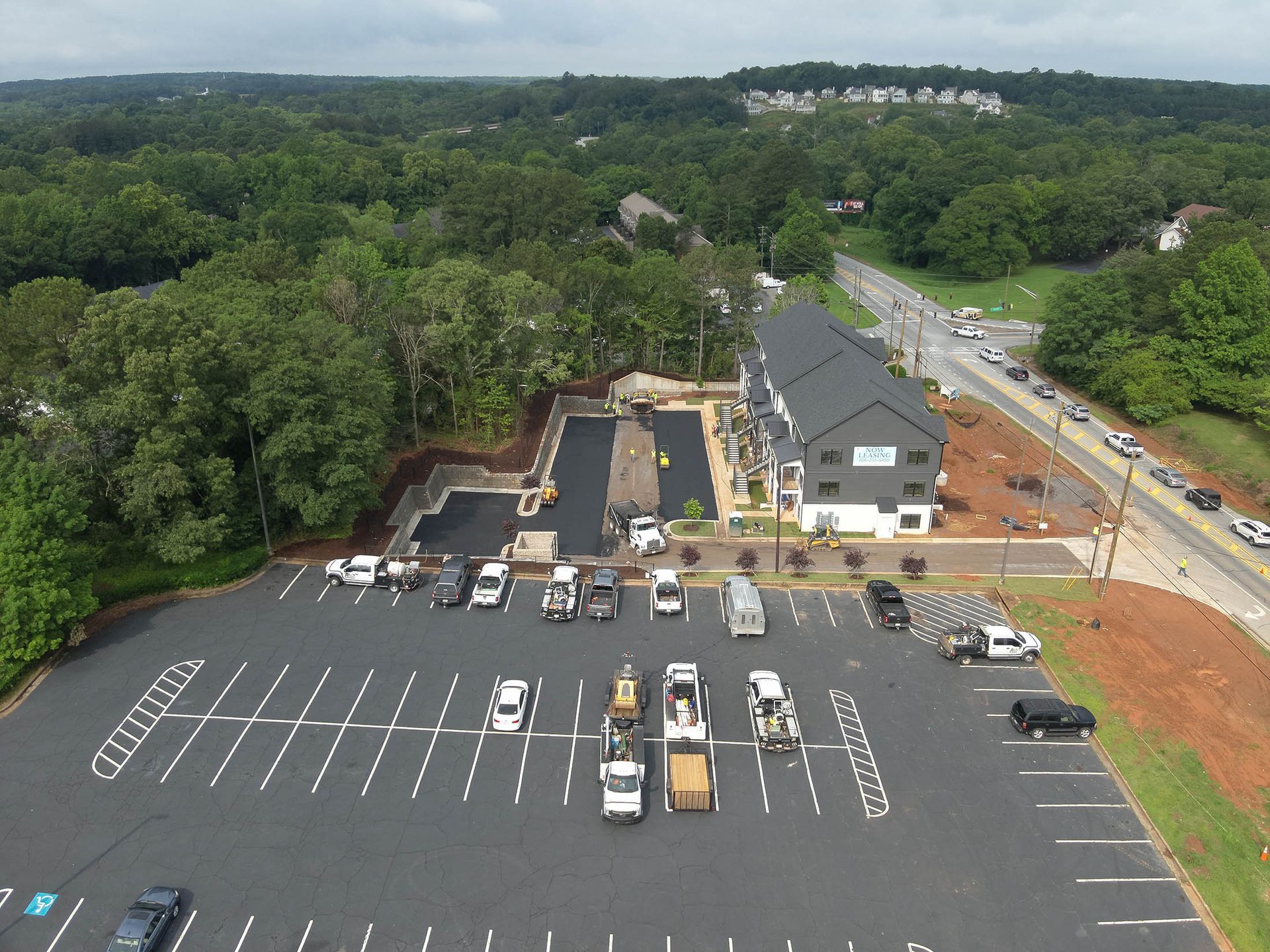 Aerial view of a parking lot, buildings, and a road surrounded by trees. Cars and trucks are present.