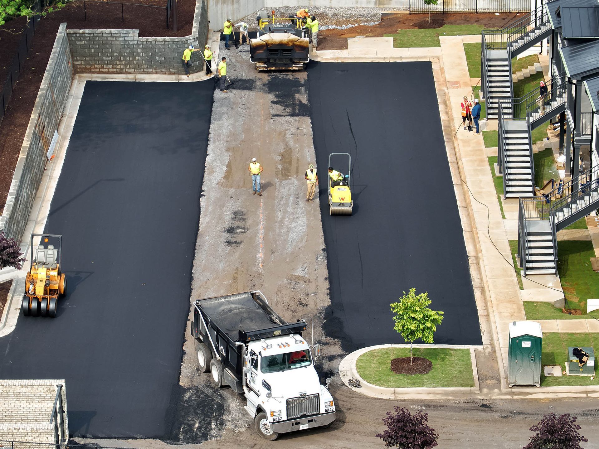 Construction workers paving a parking lot; a dump truck, steamrollers, and other machinery are present.