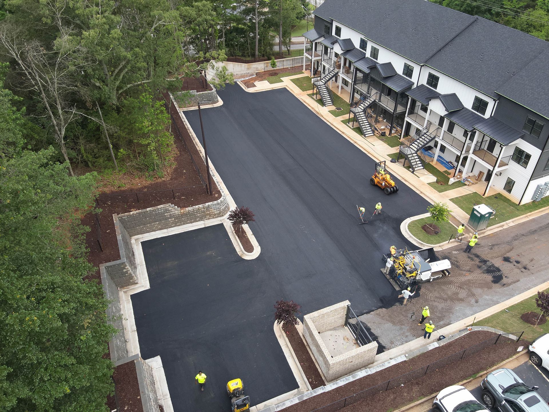 Asphalt parking lot being paved next to townhouses, workers in yellow vests, trees, and small vehicles.