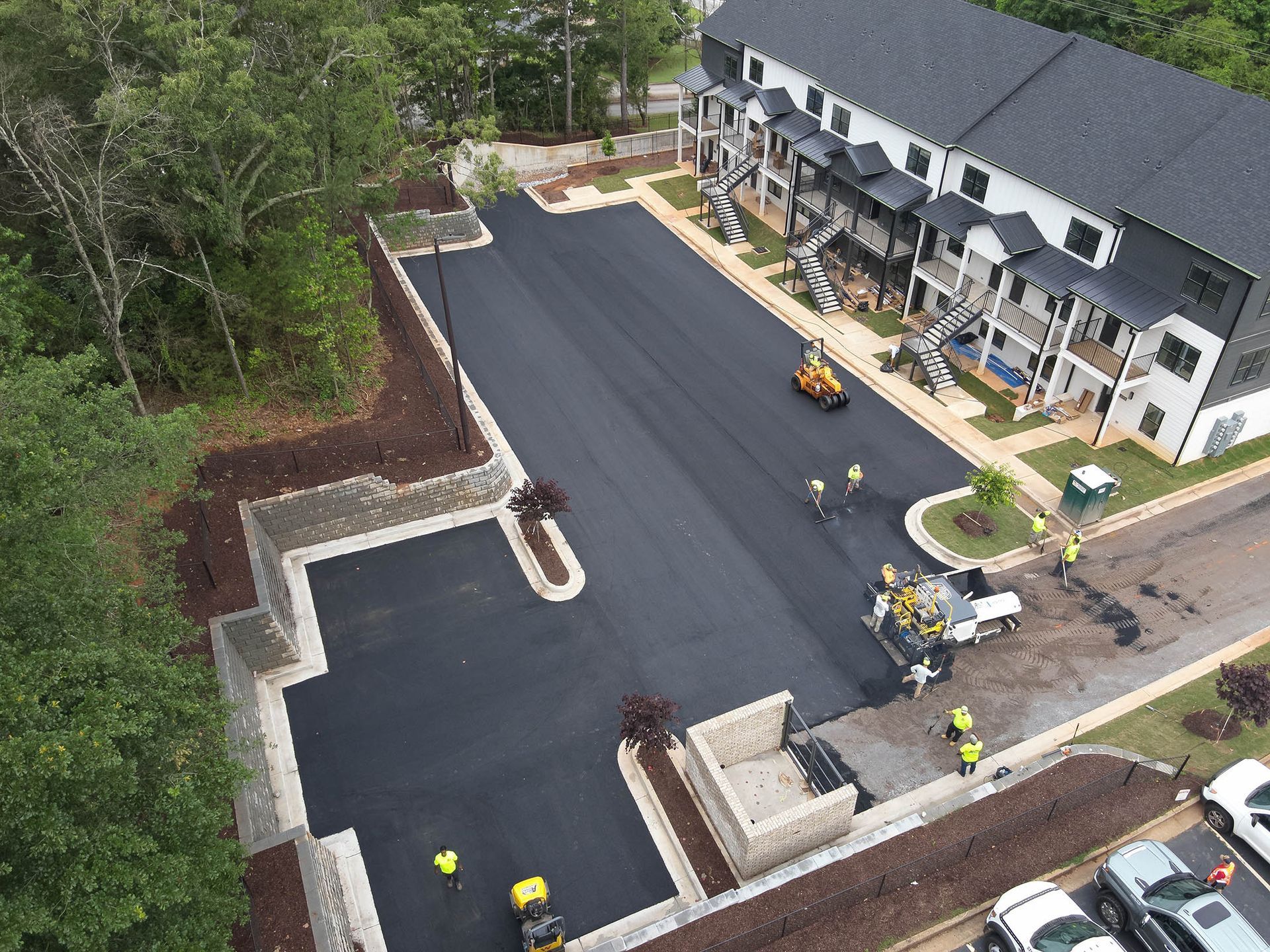 Workers paving a parking lot next to a multi-story apartment building.