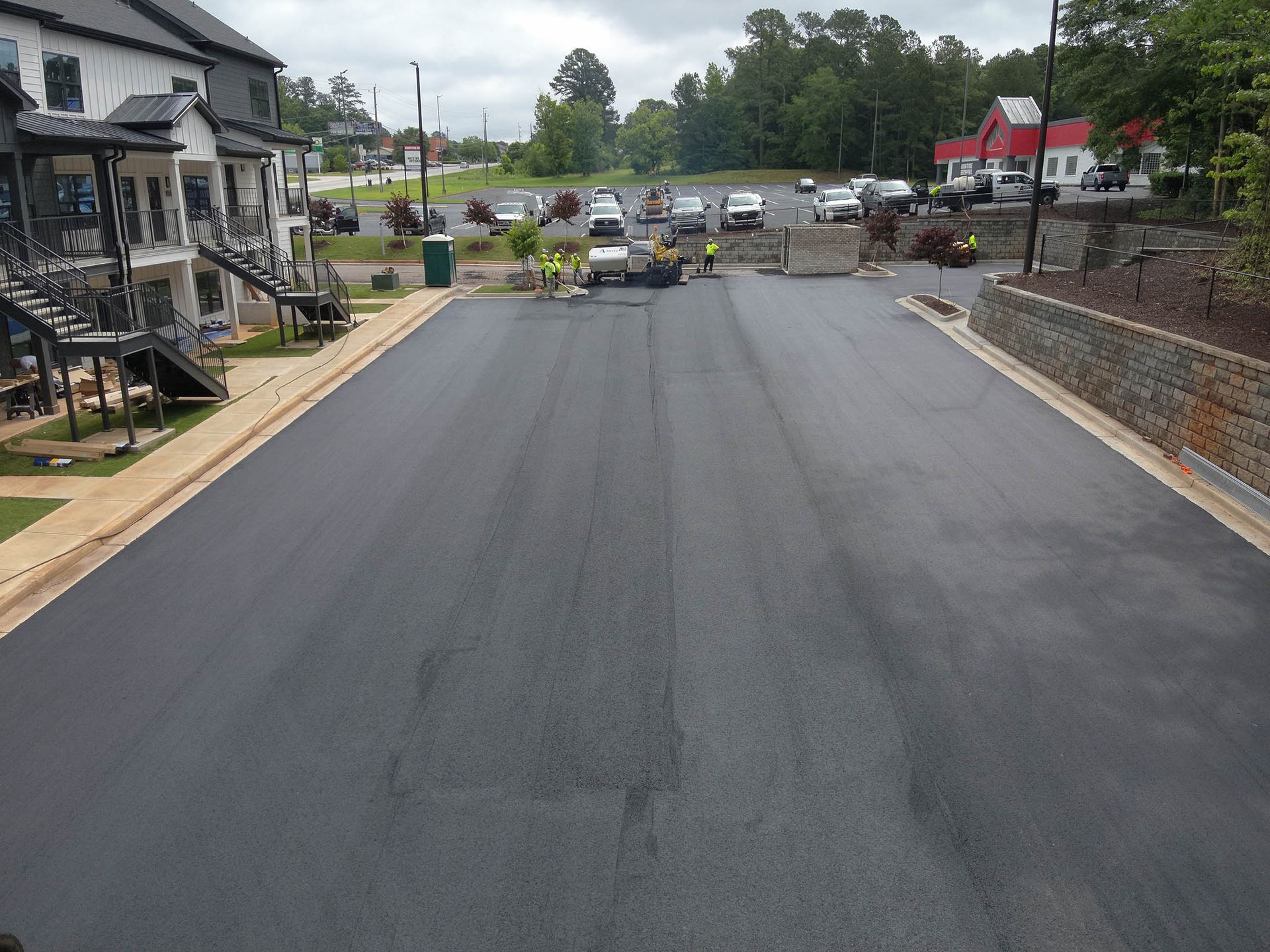 Newly paved road with workers, flanked by buildings and a stone wall. Cloudy sky.