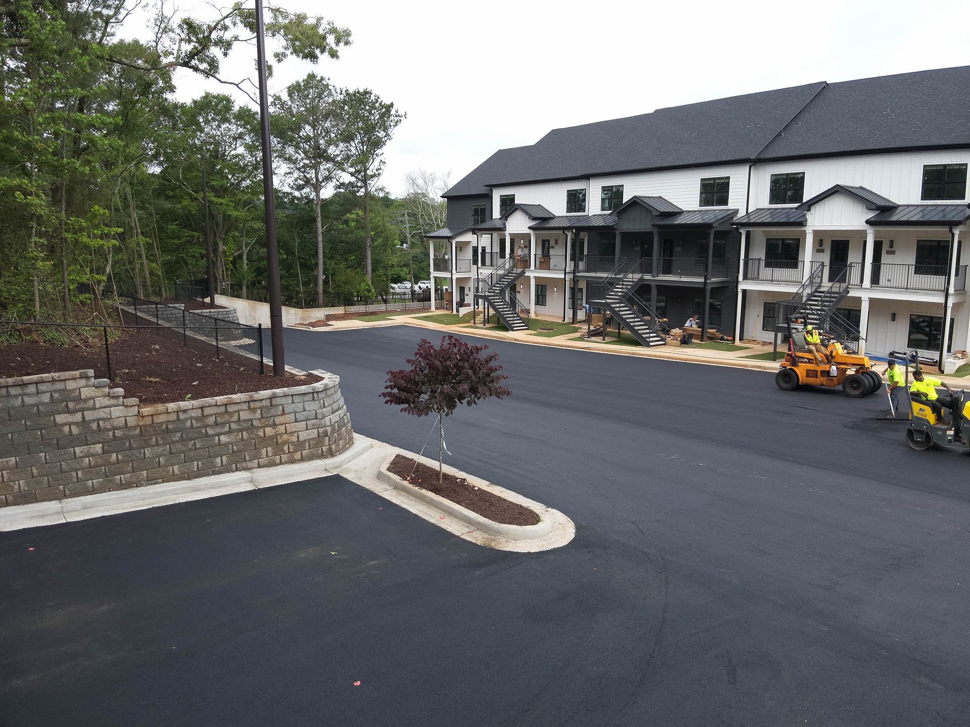 Newly paved parking lot in front of a multi-story building, with trees and construction vehicles visible.