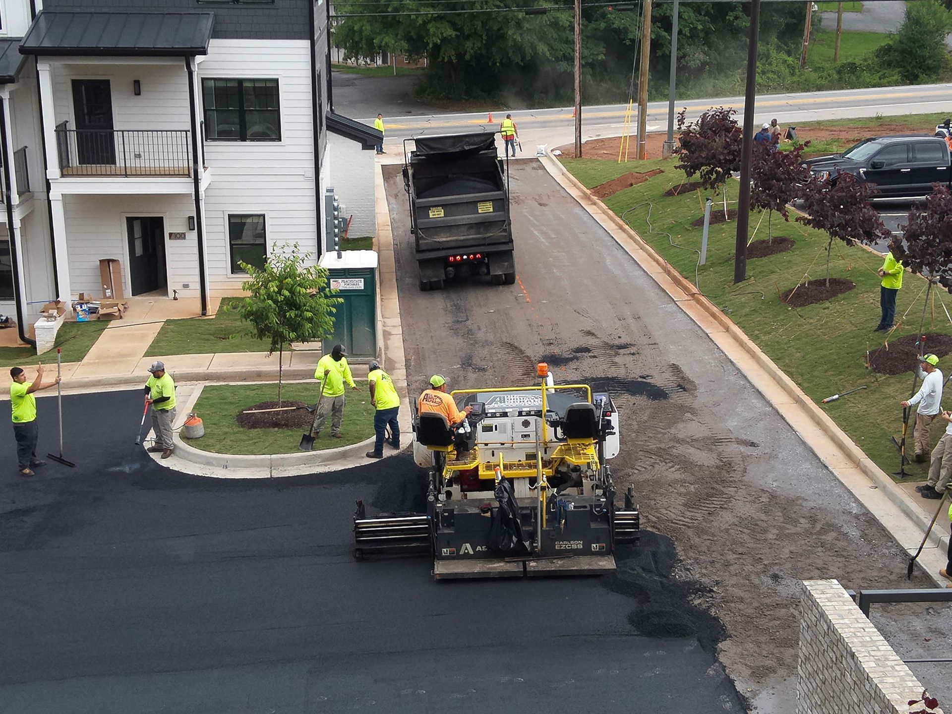 Road paving in progress. Workers in neon vests, asphalt machine, dump truck, new townhouses in the background.