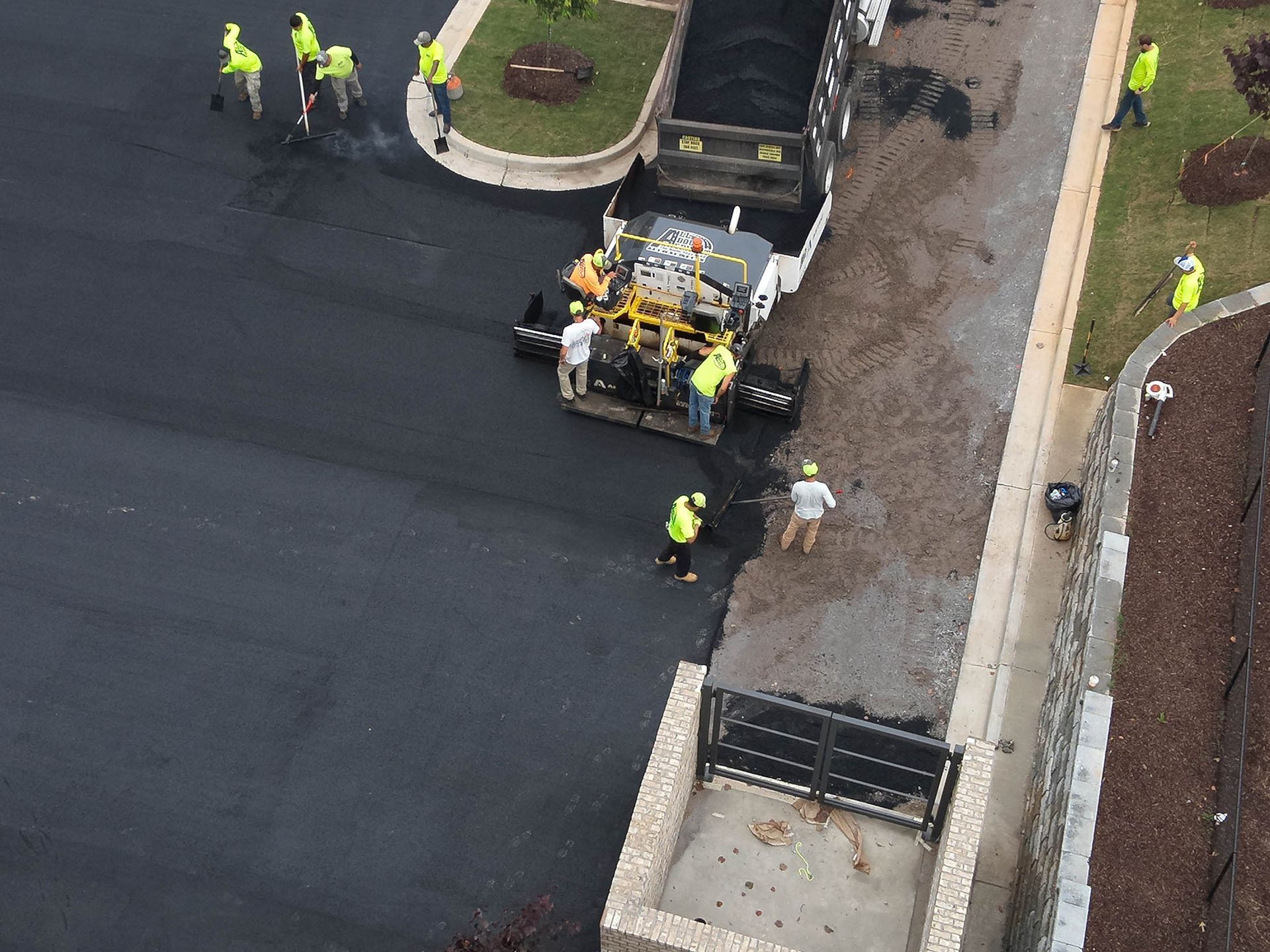 Asphalt paving crew at work, laying down new asphalt on a road near a gated entrance.