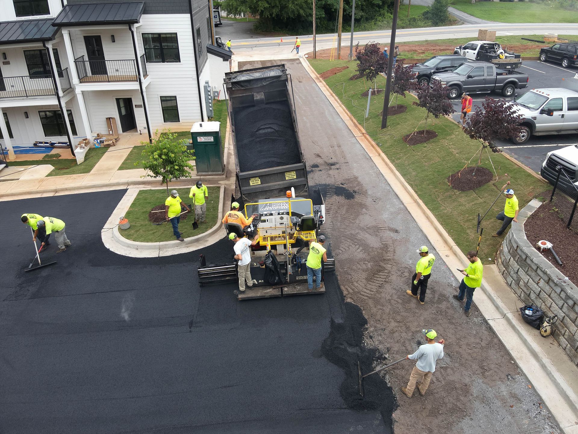 Asphalt paving crew at work, truck dumping asphalt into paver. Crew members in safety vests, apartment building in background.