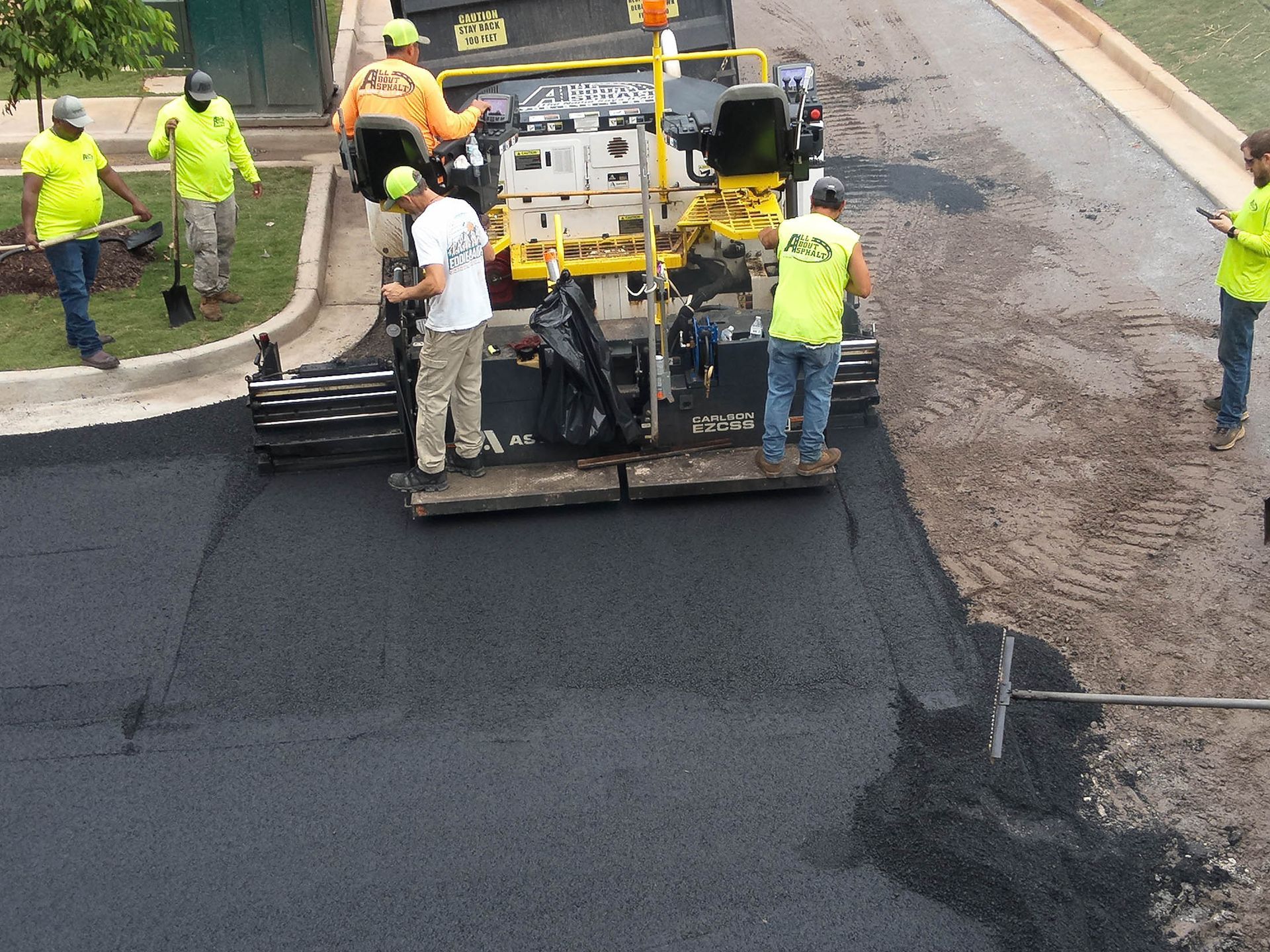 Road crew paving asphalt, with asphalt spreader and workers wearing safety vests.