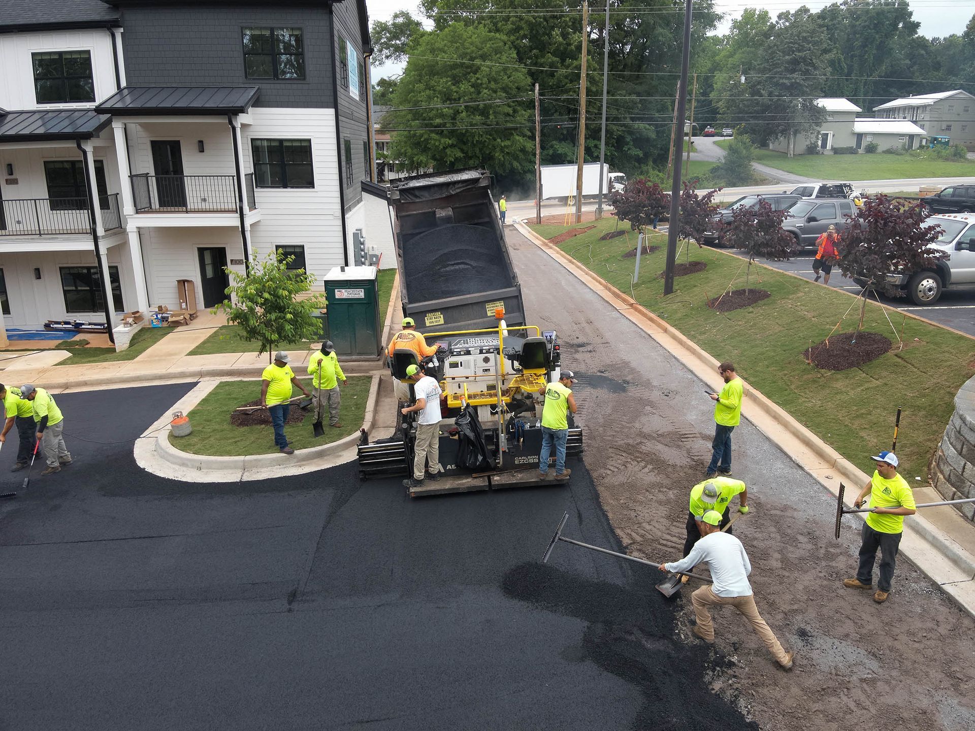 Construction crew paving a road with asphalt. Workers in yellow vests operate machinery and spread asphalt near apartment building.