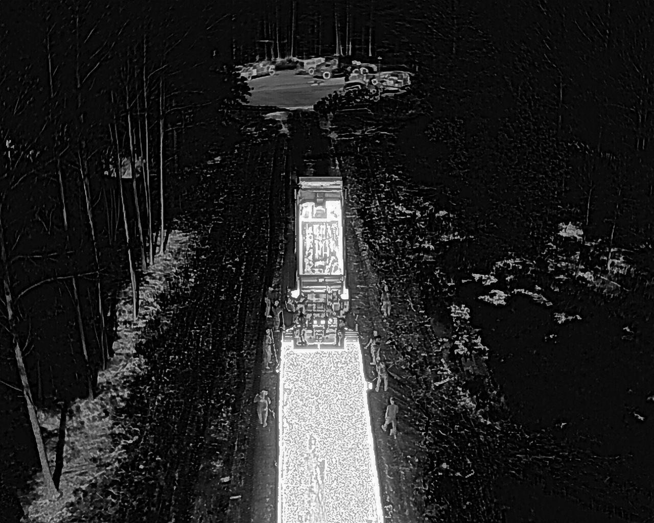 Truck driving down a dark road, surrounded by trees. Road is lit up.