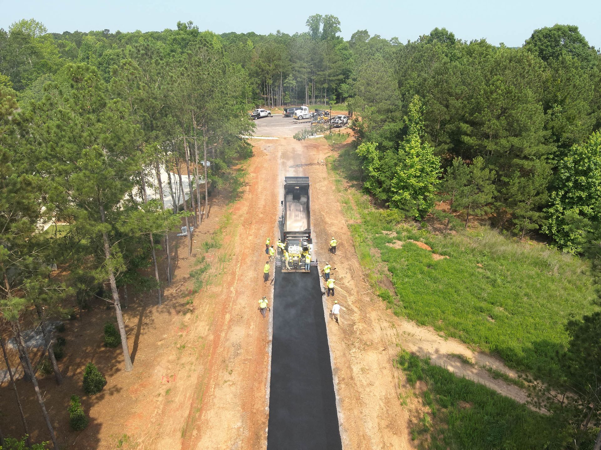 Road paving in progress, asphalt being laid by machine, workers nearby, surrounded by trees.