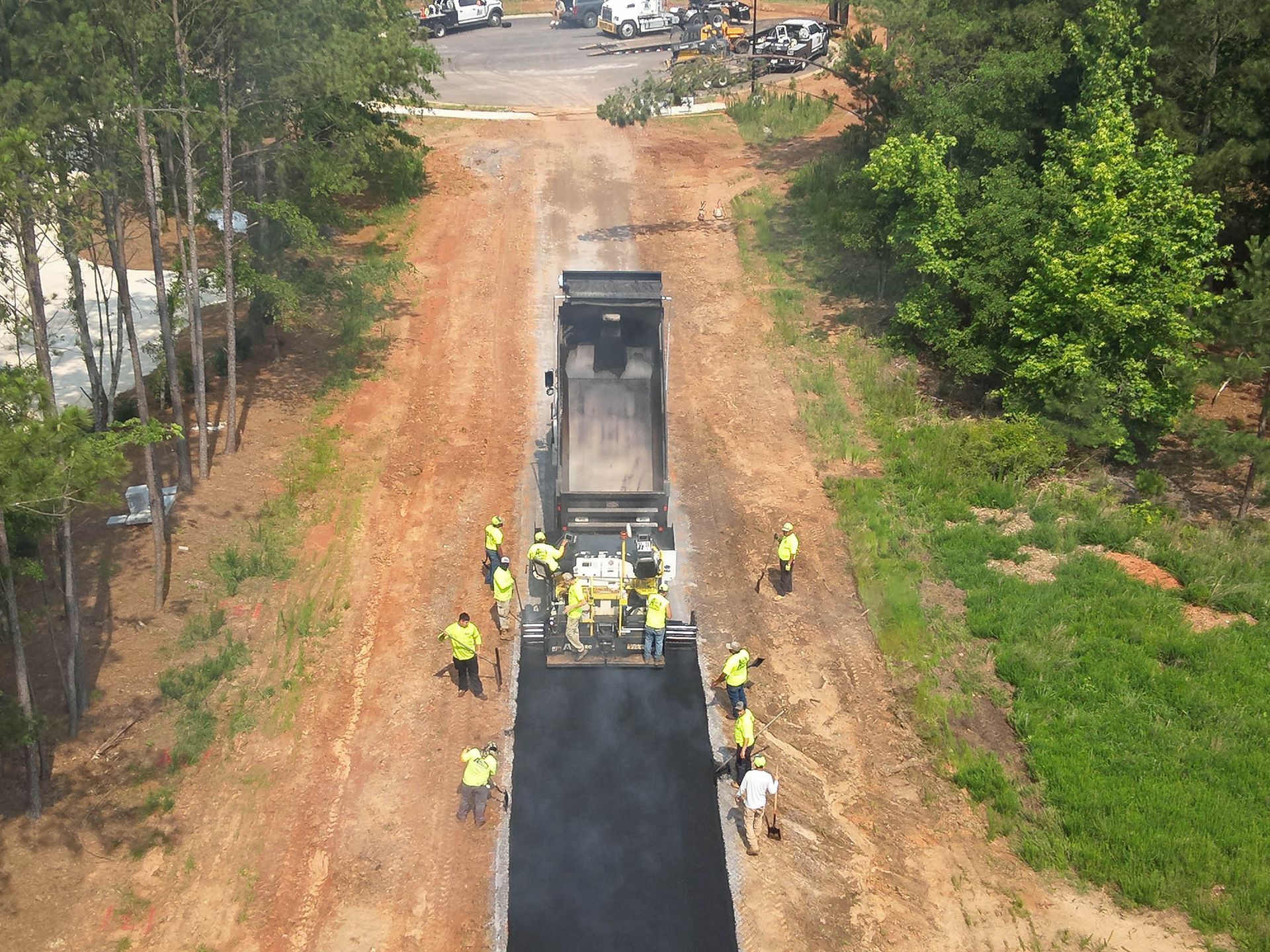 Asphalt paving in progress on a road. Workers in safety vests; truck, equipment, trees.