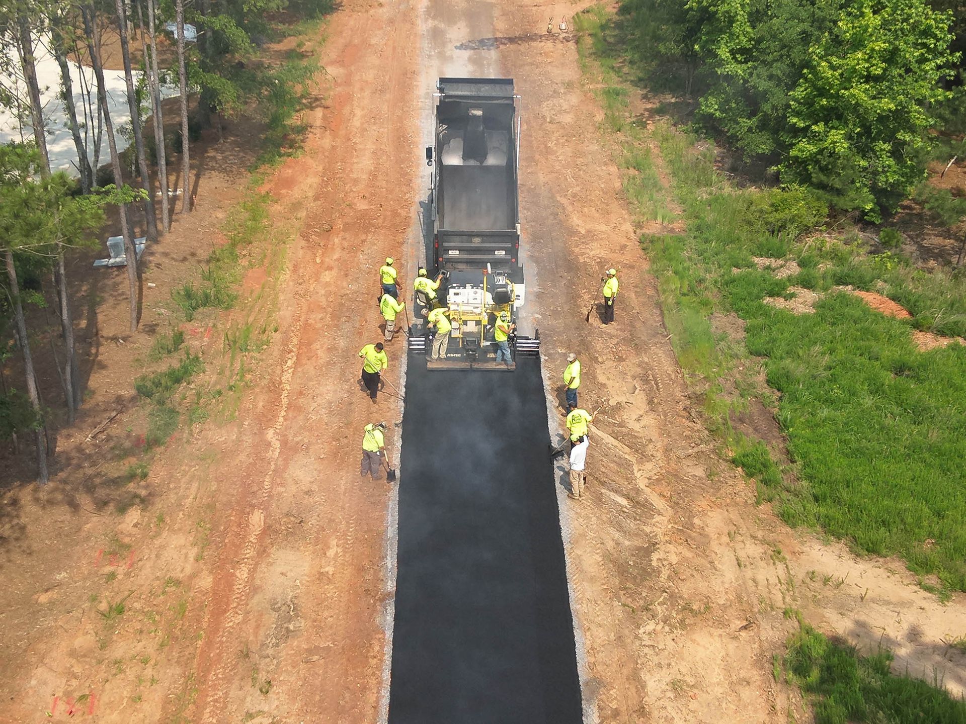 Road paving in progress; asphalt paver laying blacktop with workers in safety vests; dirt road setting.