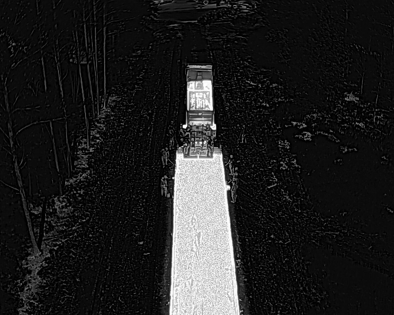 Overhead view of a narrow, bright bridge. A vehicle with a tall cabin crosses a dark body of water.