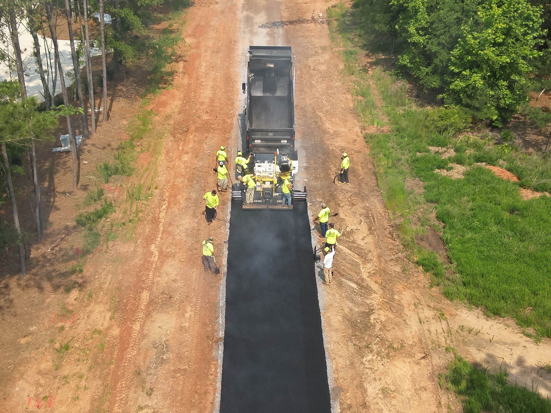 Road construction. Asphalt paver laying fresh pavement, workers in yellow vests, rural setting.