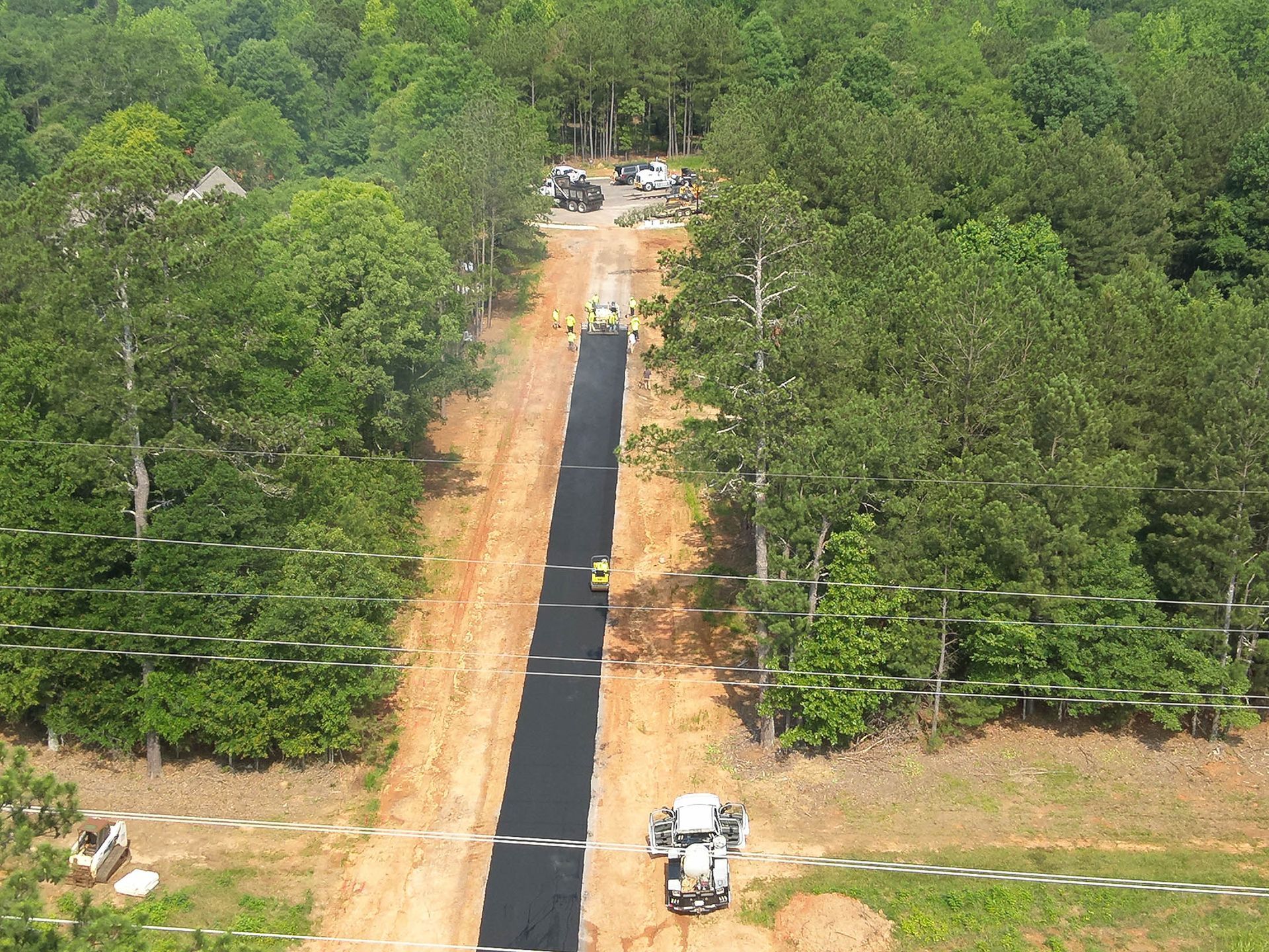 Aerial view of a road construction site in a wooded area. Black asphalt is laid down, surrounded by dirt and trees.