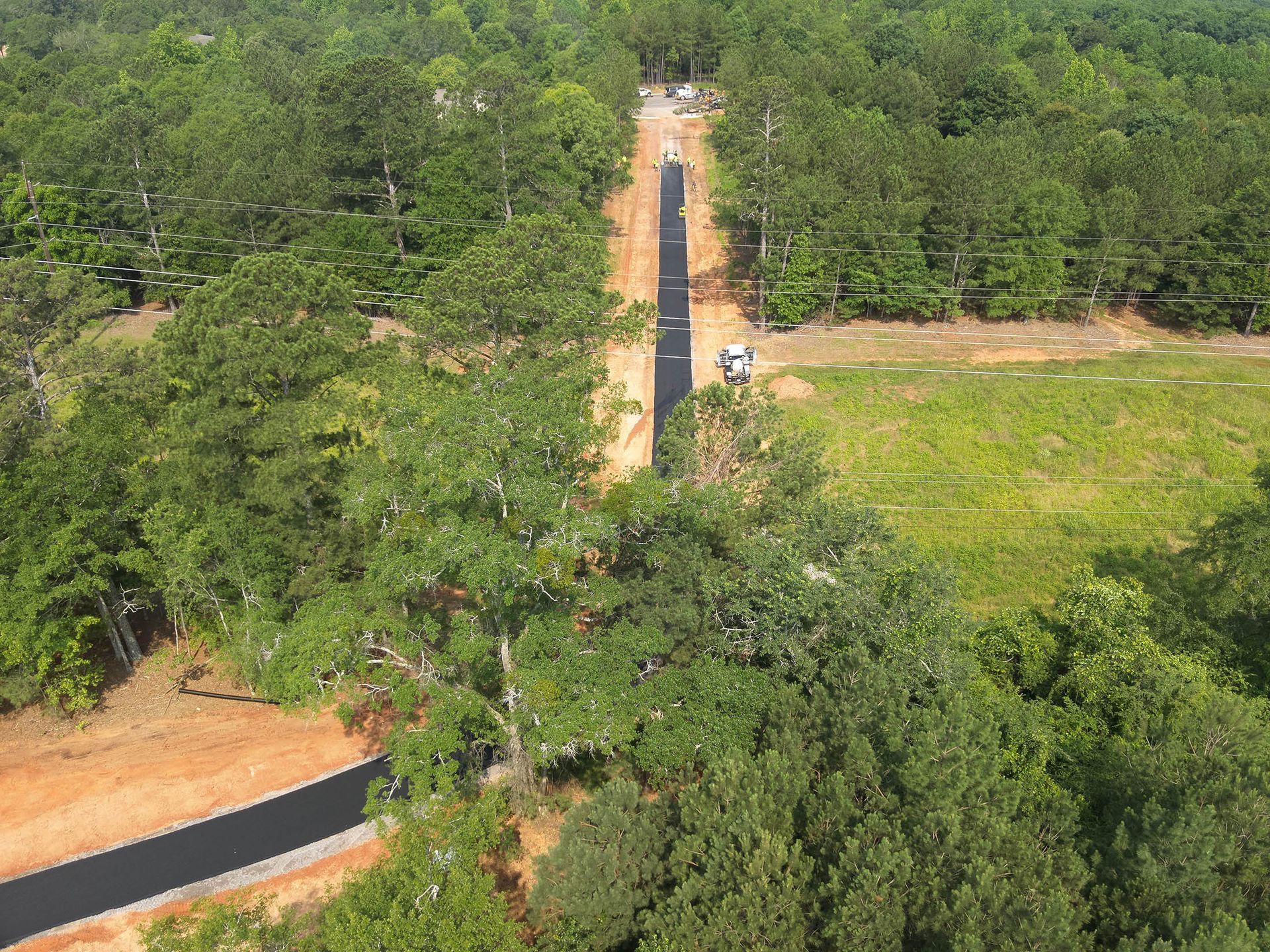 Asphalt road cuts through a green forest, leading to clearing and construction site.