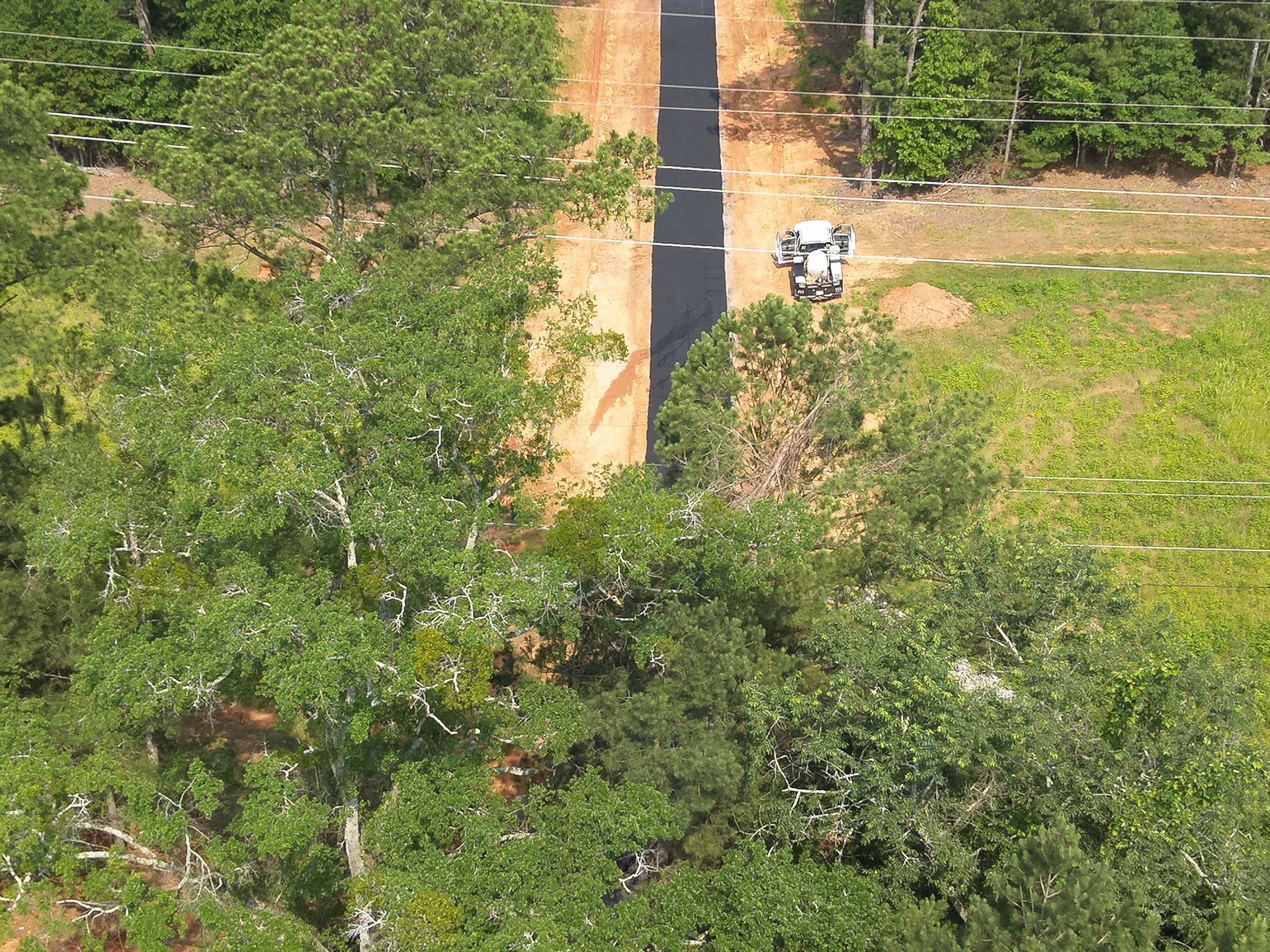 Road construction in a forest. A dark paved road extends between trees, a work vehicle sits at a clearing.