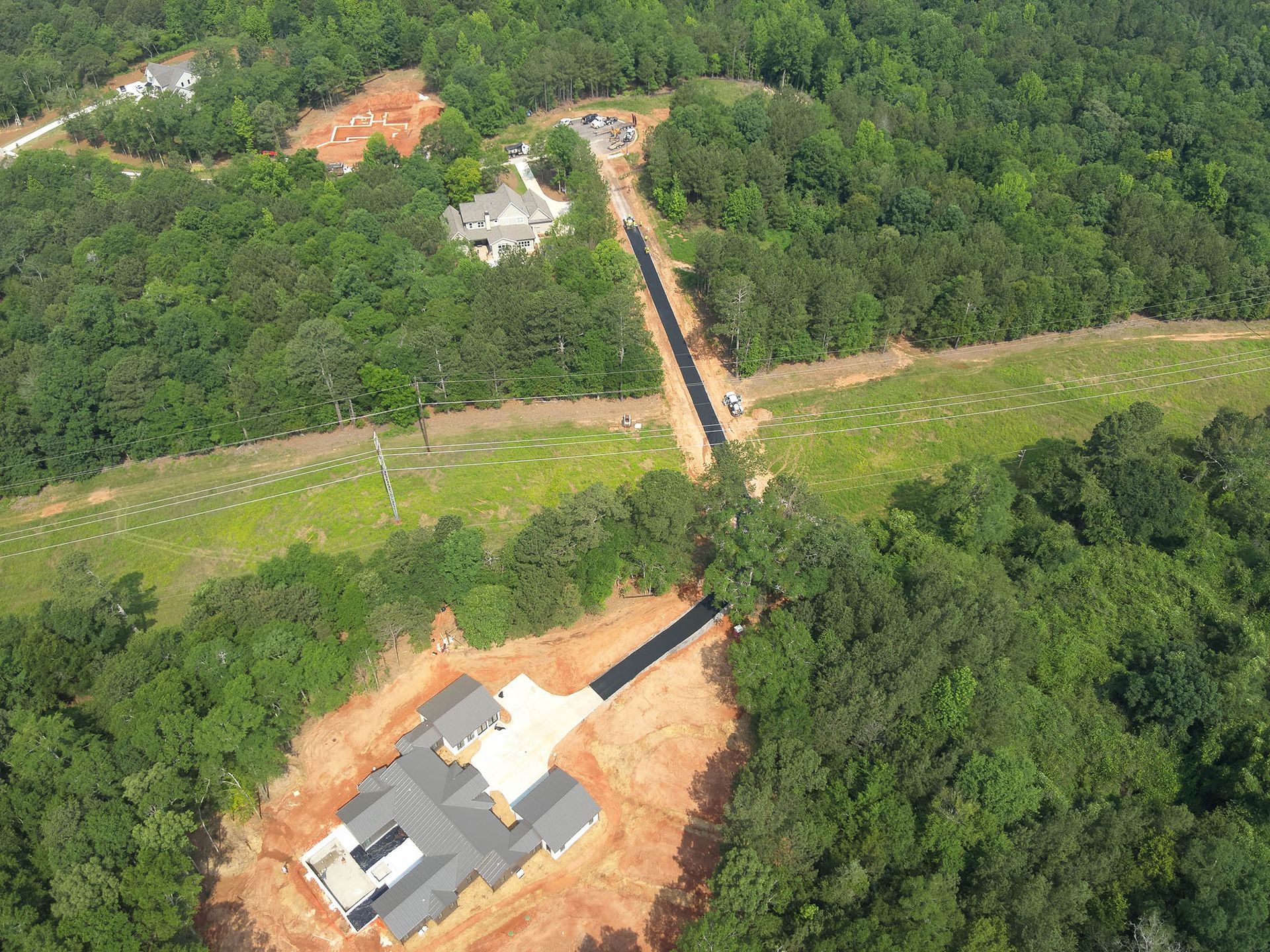 Aerial view of a construction site. A new house under construction sits in a cleared area, with a long road.