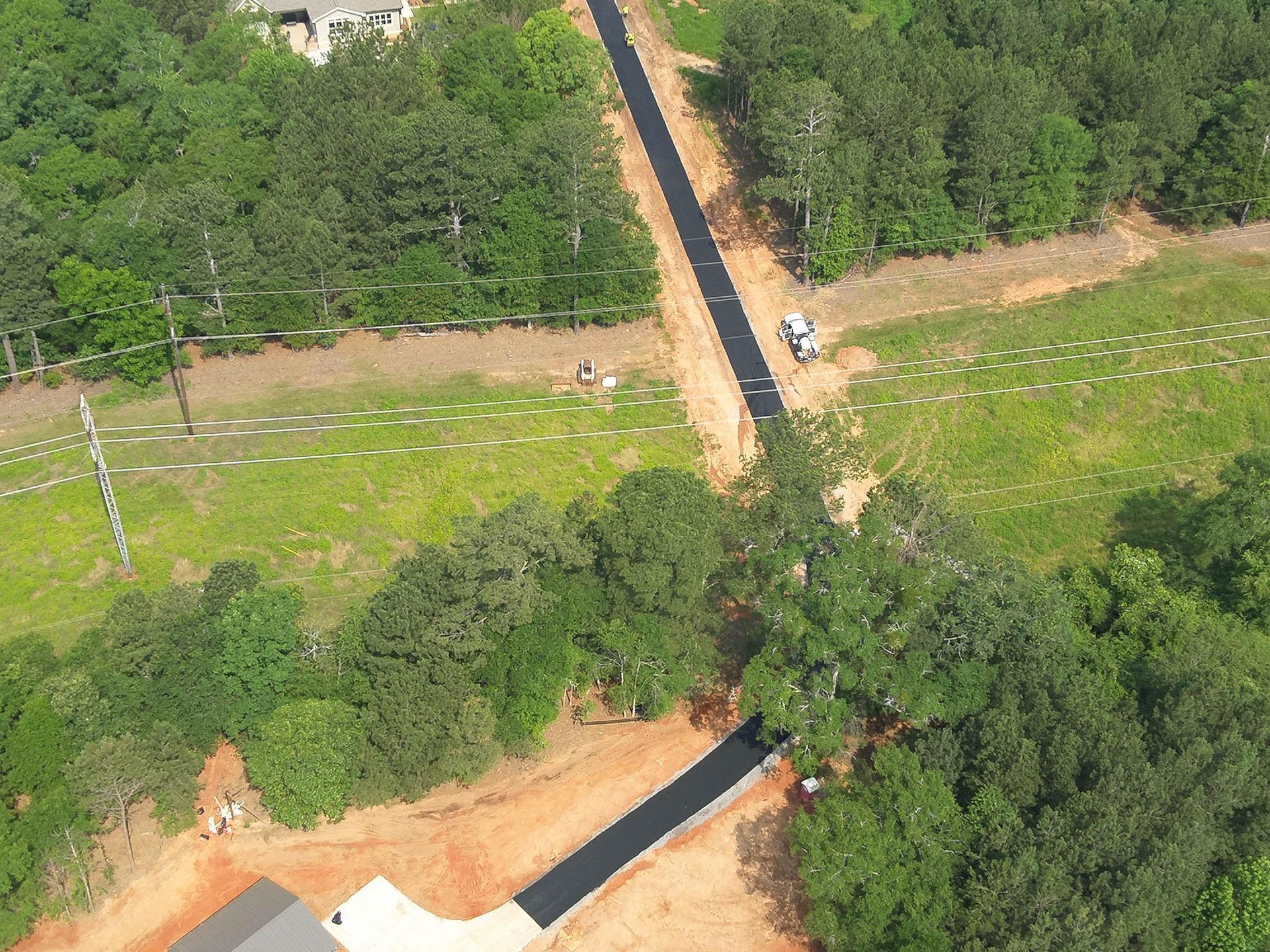 Aerial view of a newly paved road winding through a wooded area. A vehicle is at work. Green grass and trees.