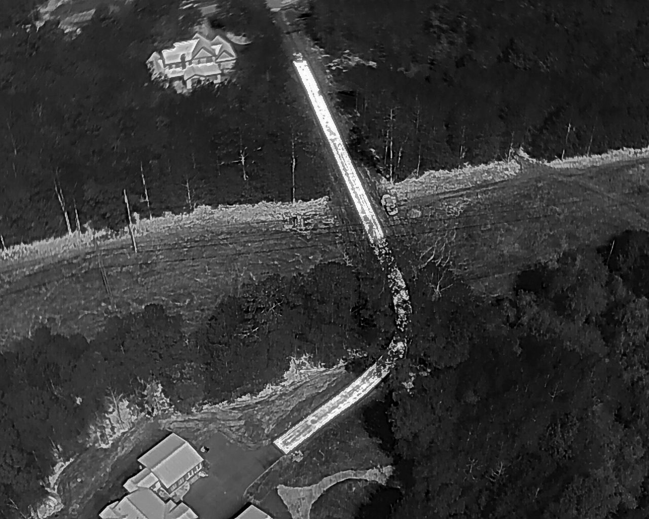 Aerial view of a road cutting through a forest. A house is at the top, and some buildings are at the bottom.