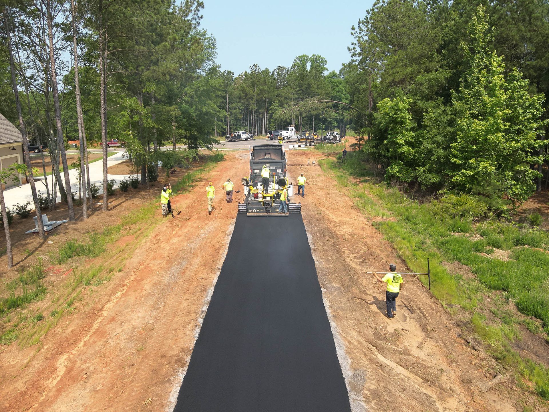 Road construction. Asphalt being laid by a truck and crew, surrounded by dirt, trees, and workers.