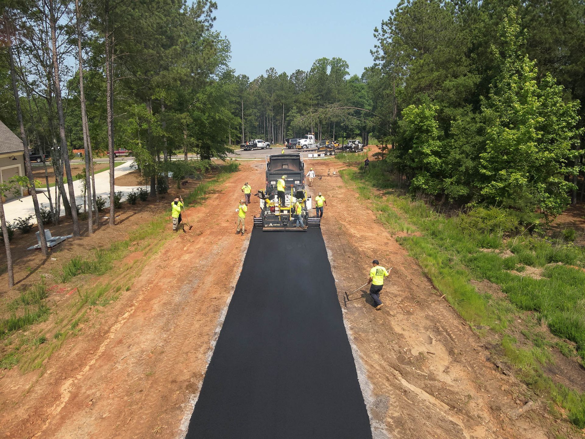 Road paving in progress. Workers in yellow vests operate machinery and spread asphalt on a dirt road lined with trees.