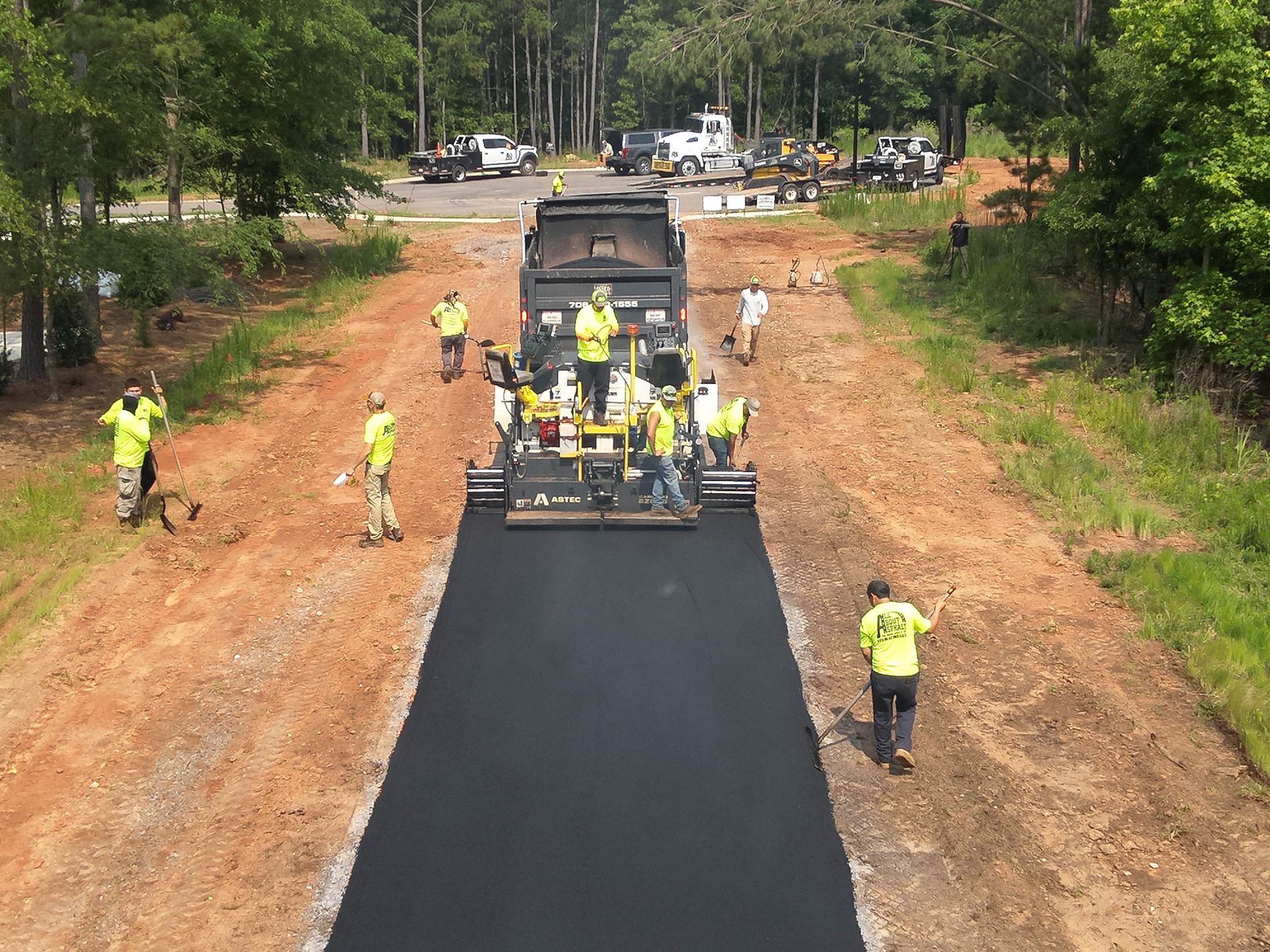 Workers in safety vests paving a road with an asphalt paver. Dirt and trees in the background.