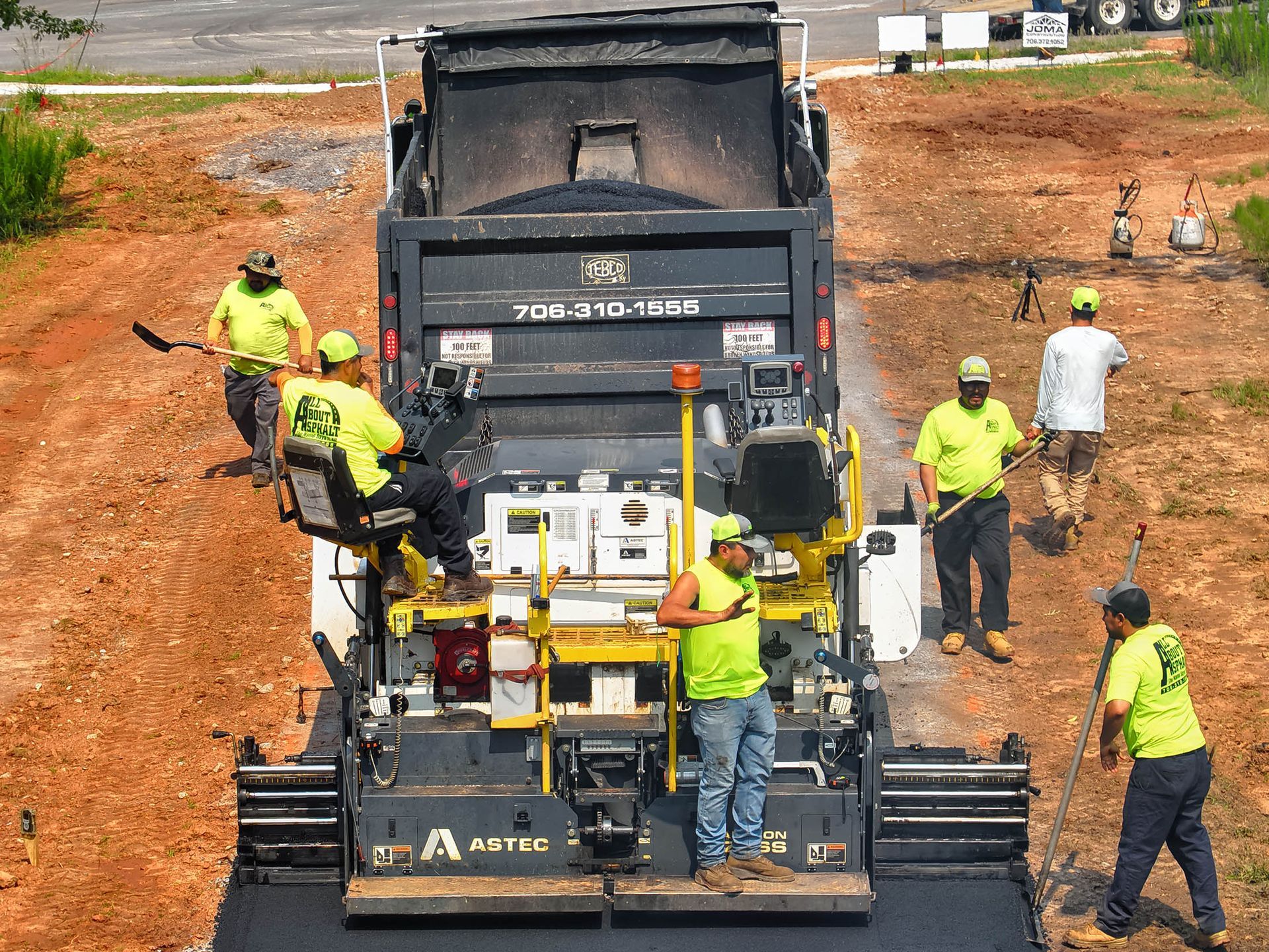 Road paving crew operating a large asphalt machine, laying down new pavement. Workers in neon green vests, outdoors.