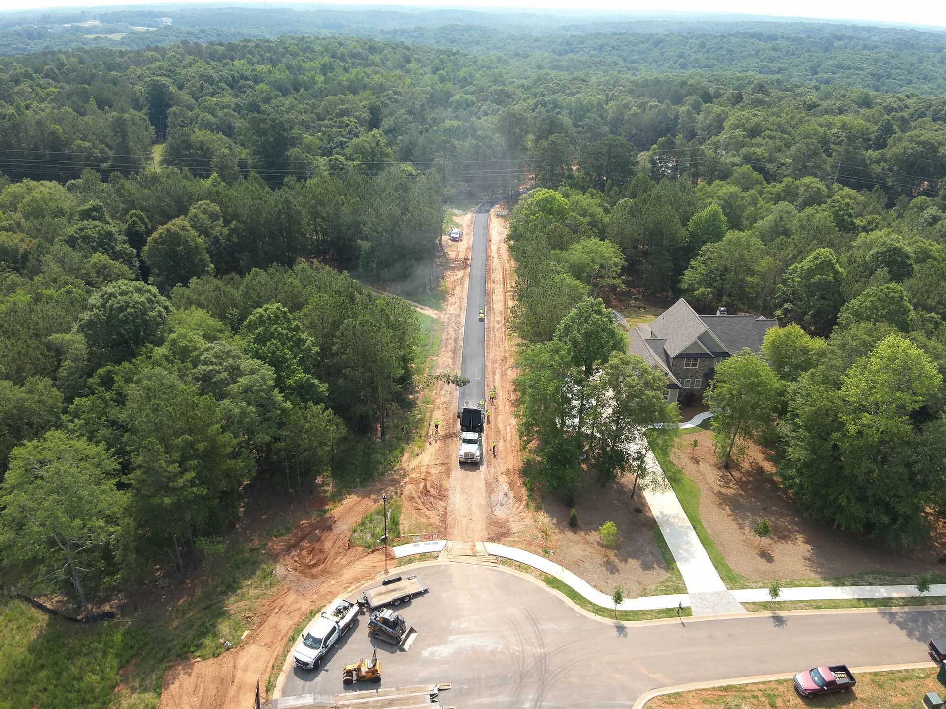Aerial view of road construction through a forest, with a house visible on the right.