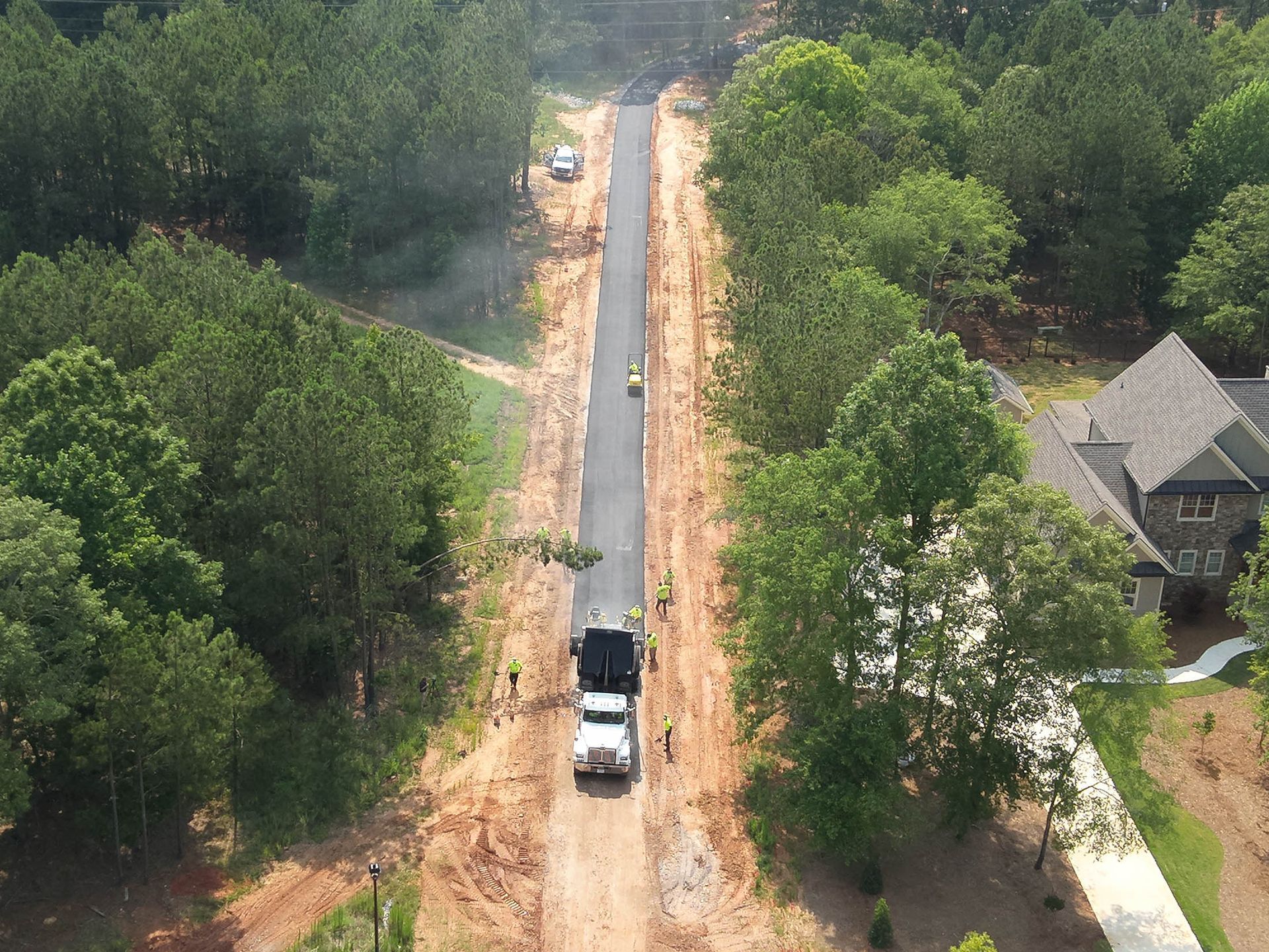 Road paving in progress, surrounded by trees and a house. A dump truck is at work on the new road.
