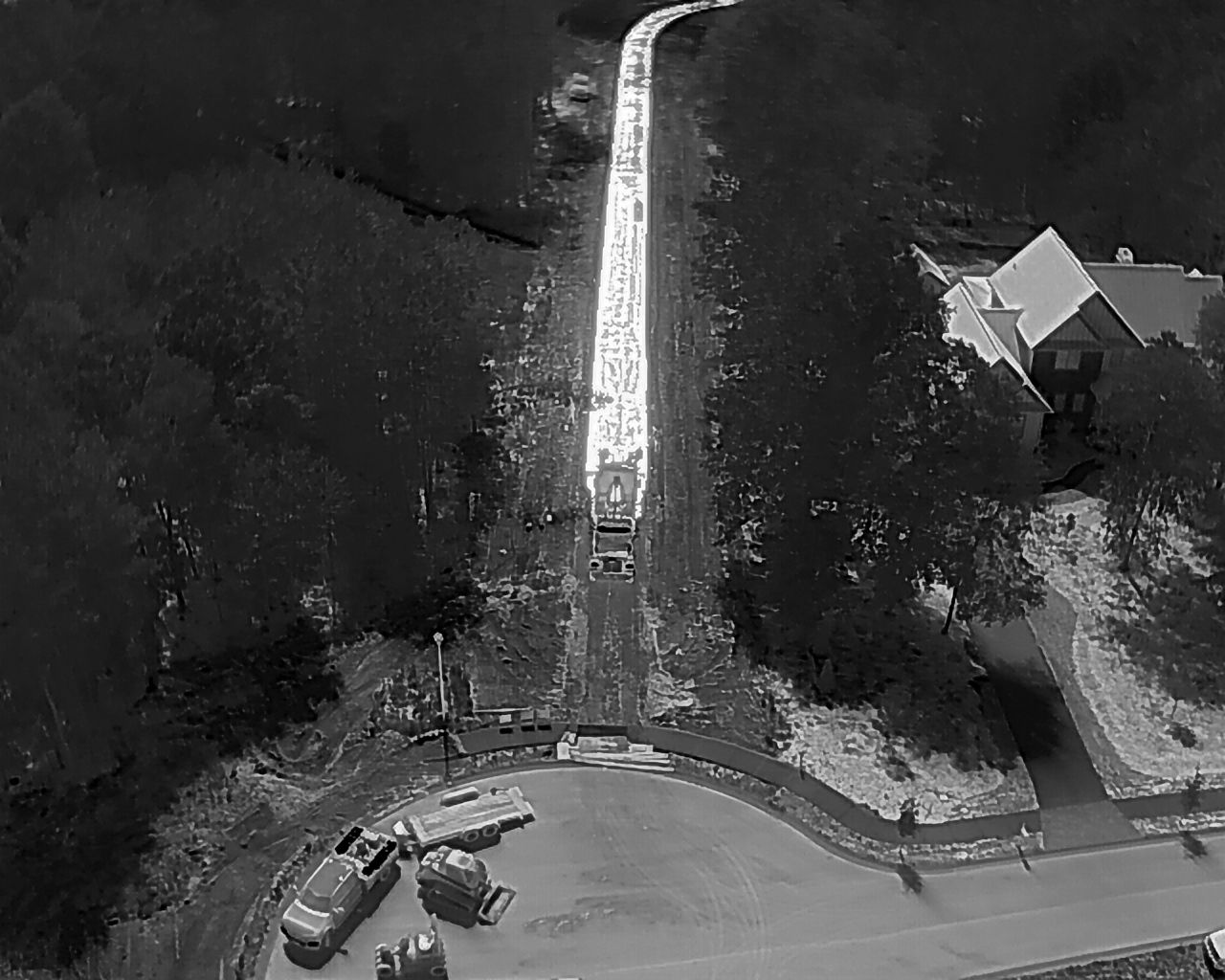 Aerial view of a road leading through a forest with a house on the right, vehicles in the lower left.