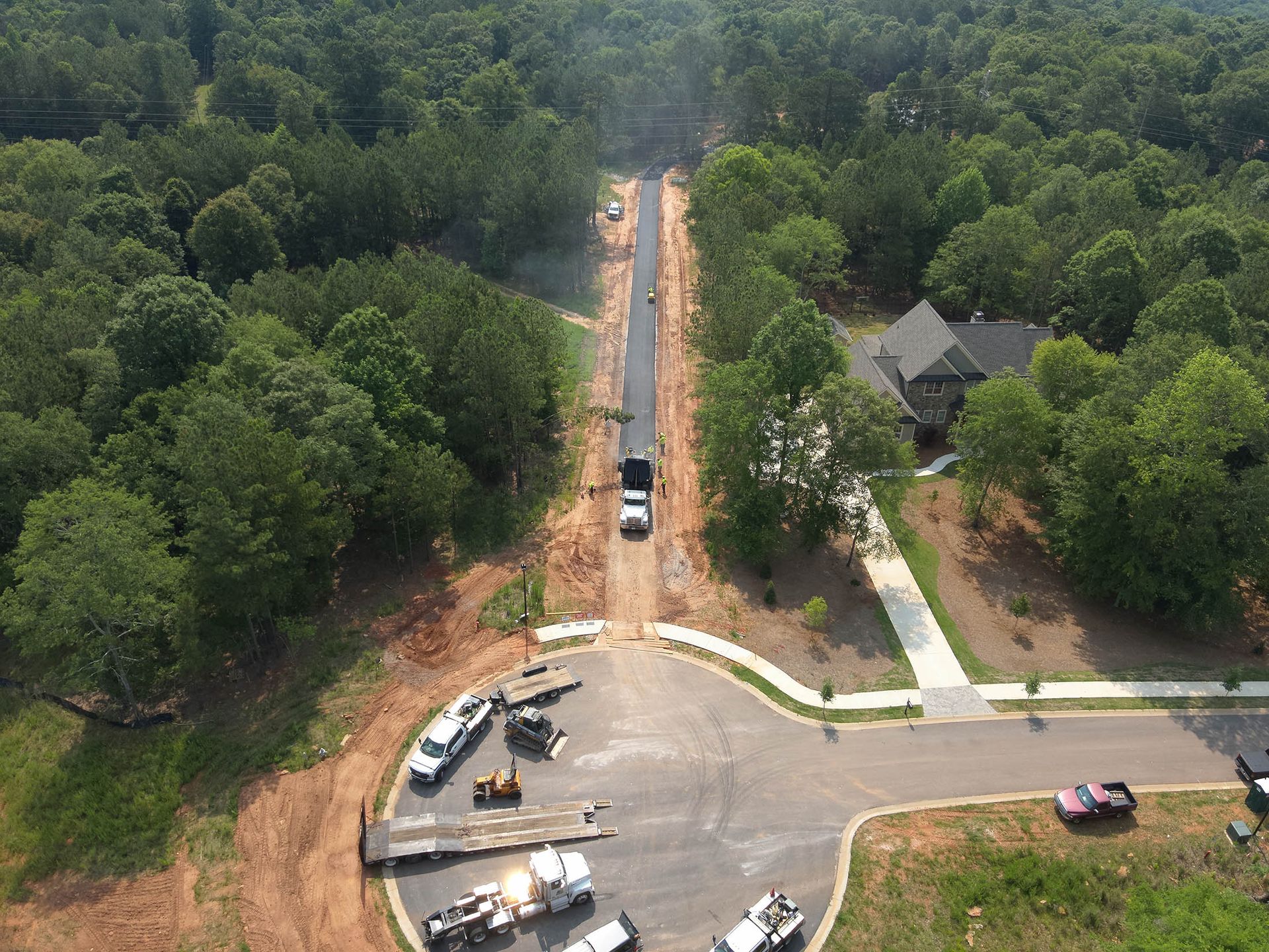Aerial view of road construction in a wooded area, with trucks and equipment visible.